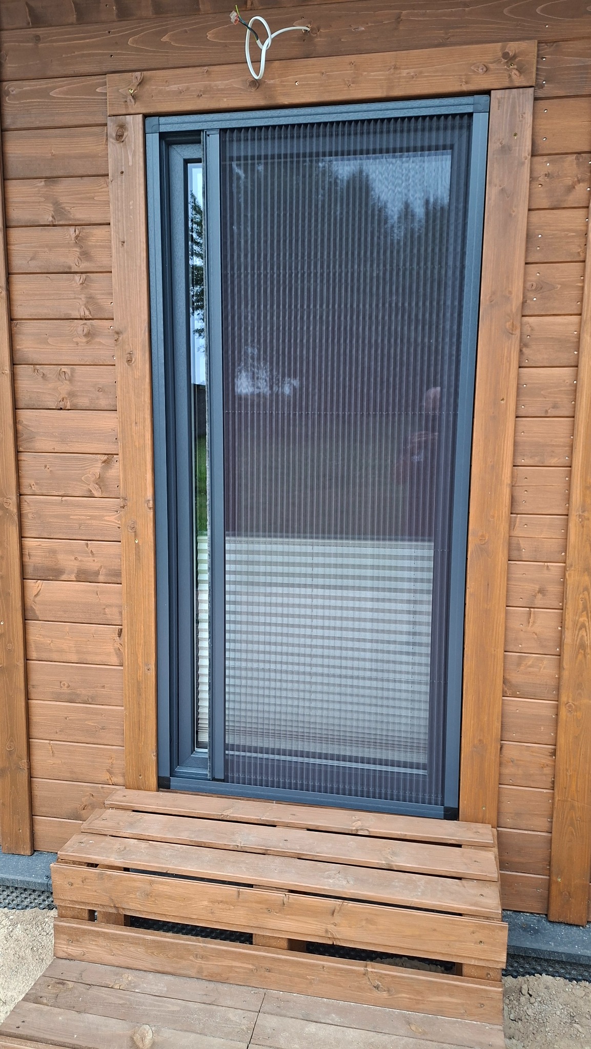 Zamontowana przesuwna moskitiera plisowana w oknie drewnianego domu, widoczna z zewnątrz, z drewnianym progiem.