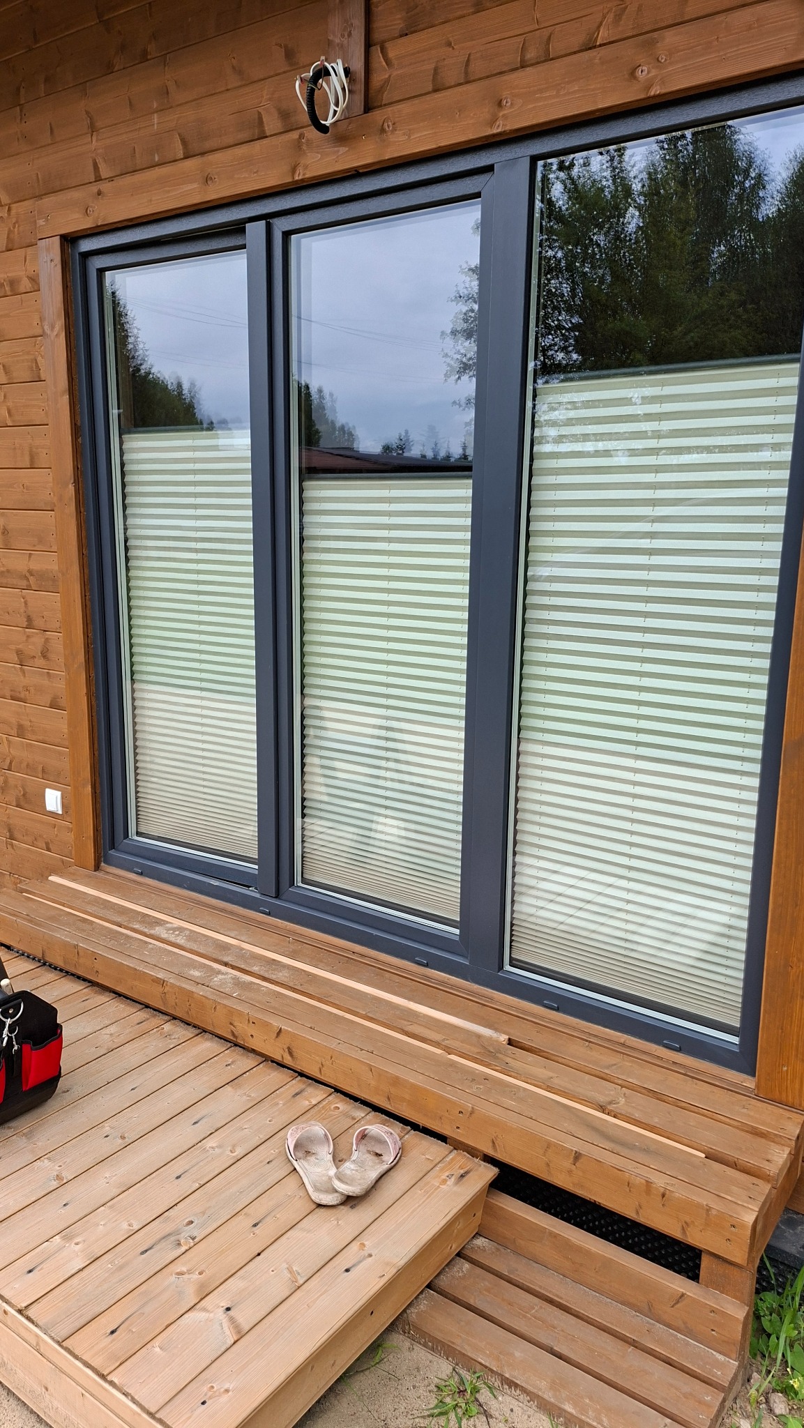 Trzy okna z szarymi ramami, osłonięte plisami w kolorze kremowym, widoczne z zewnątrz drewnianego domu. Na tarasie leżą klapki.