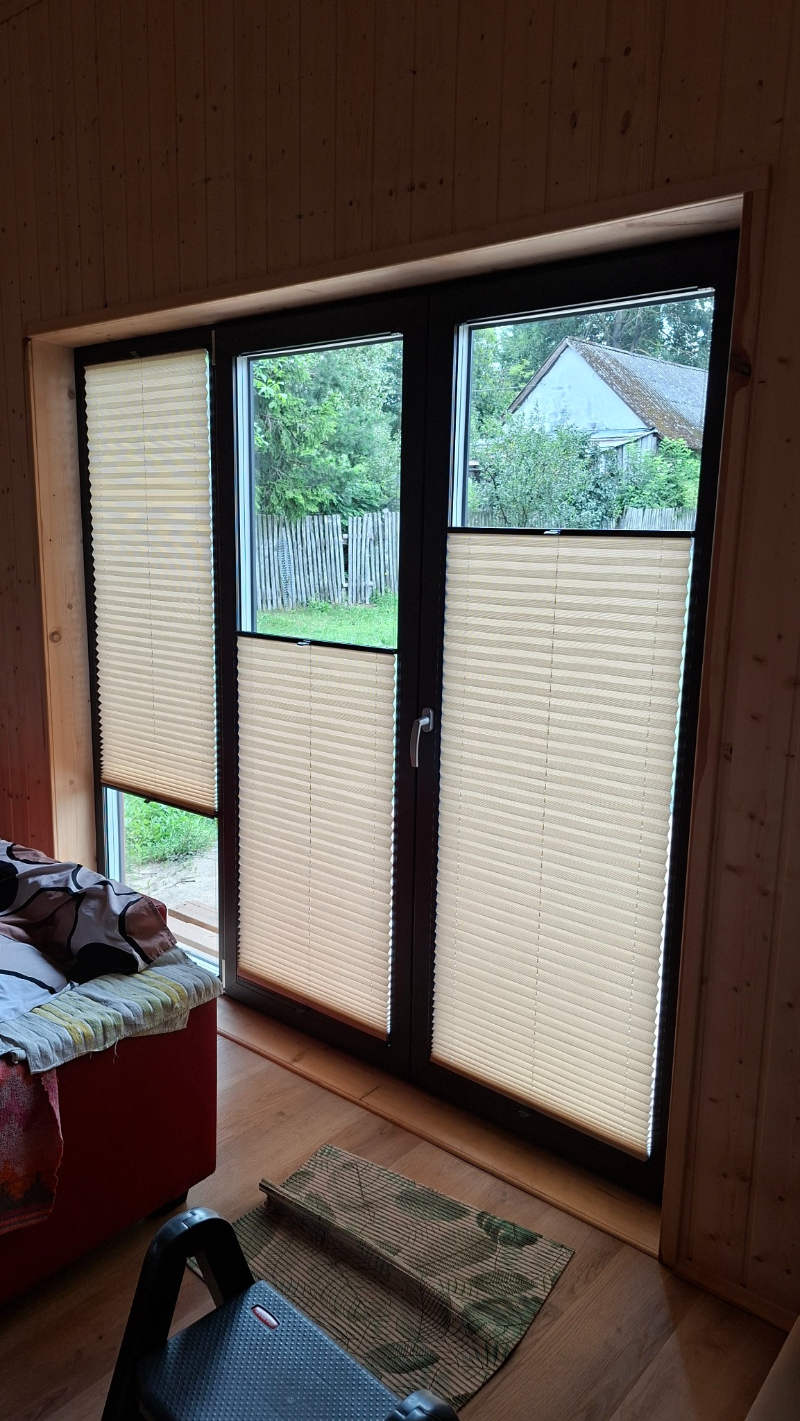Beżowe plisy okienne zamontowane w oknie z ciemną ramą w drewnianym wnętrzu. Widok na zewnątrz, gdzie widać zieleń i budynek.