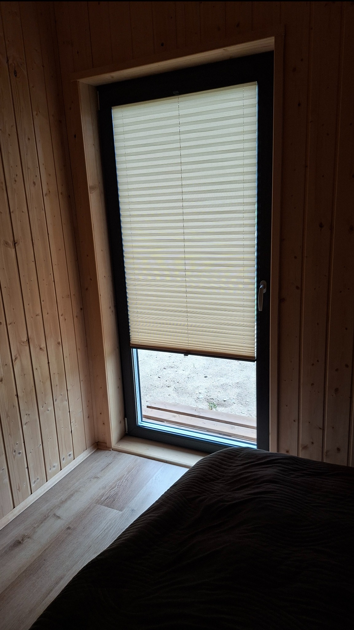 Beżowa plisa okienna zamontowana w oknie w pokoju z drewnianą boazerią. Częściowo zasłonięte okno z widokiem na zewnątrz. Widoczny fragment łóżka.