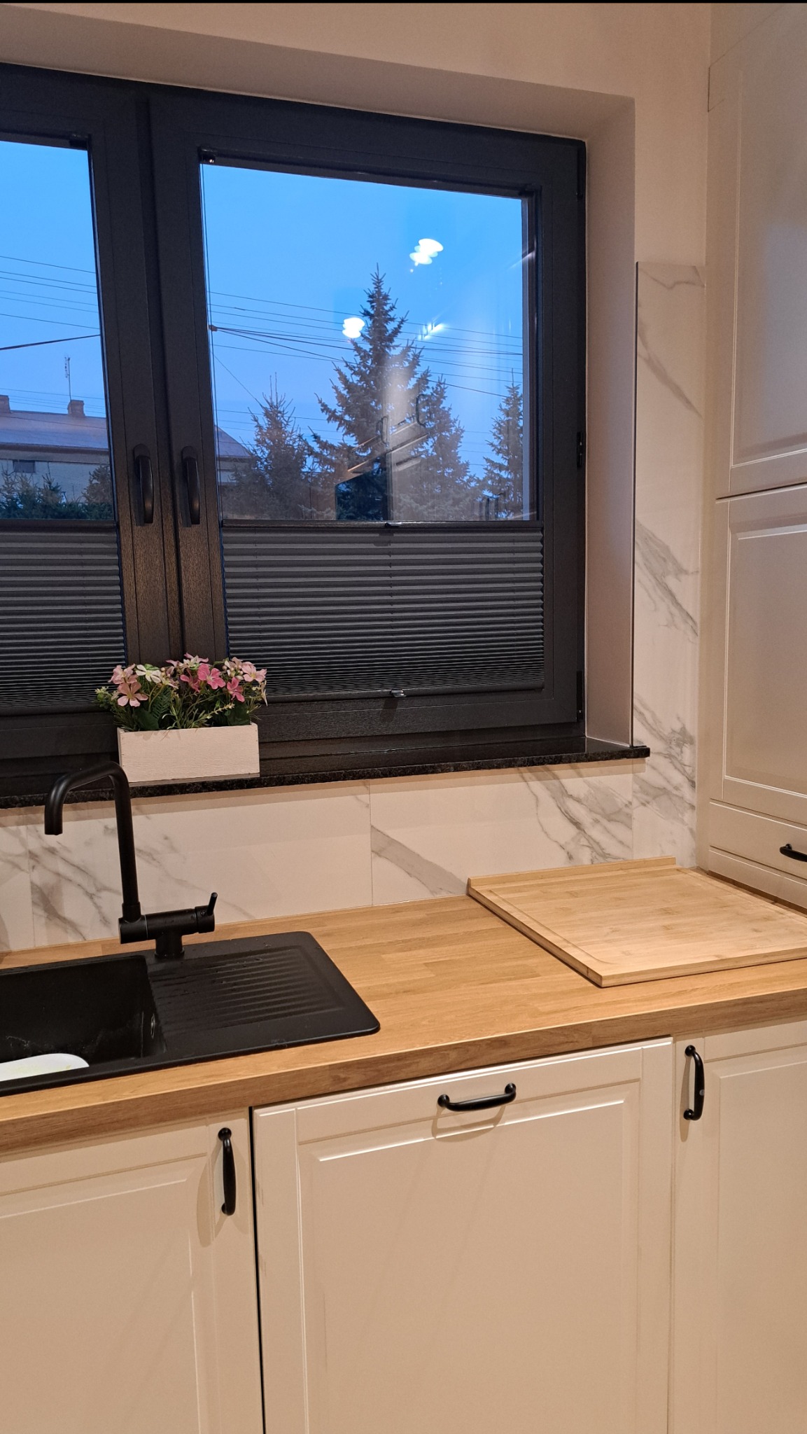 Ciemne plisy okienne w kuchni z czarnym oknem i kwiatami w doniczce na parapecie. Widok na zewnątrz z drzewami. Jasne meble kuchenne z drewnianym blatem.