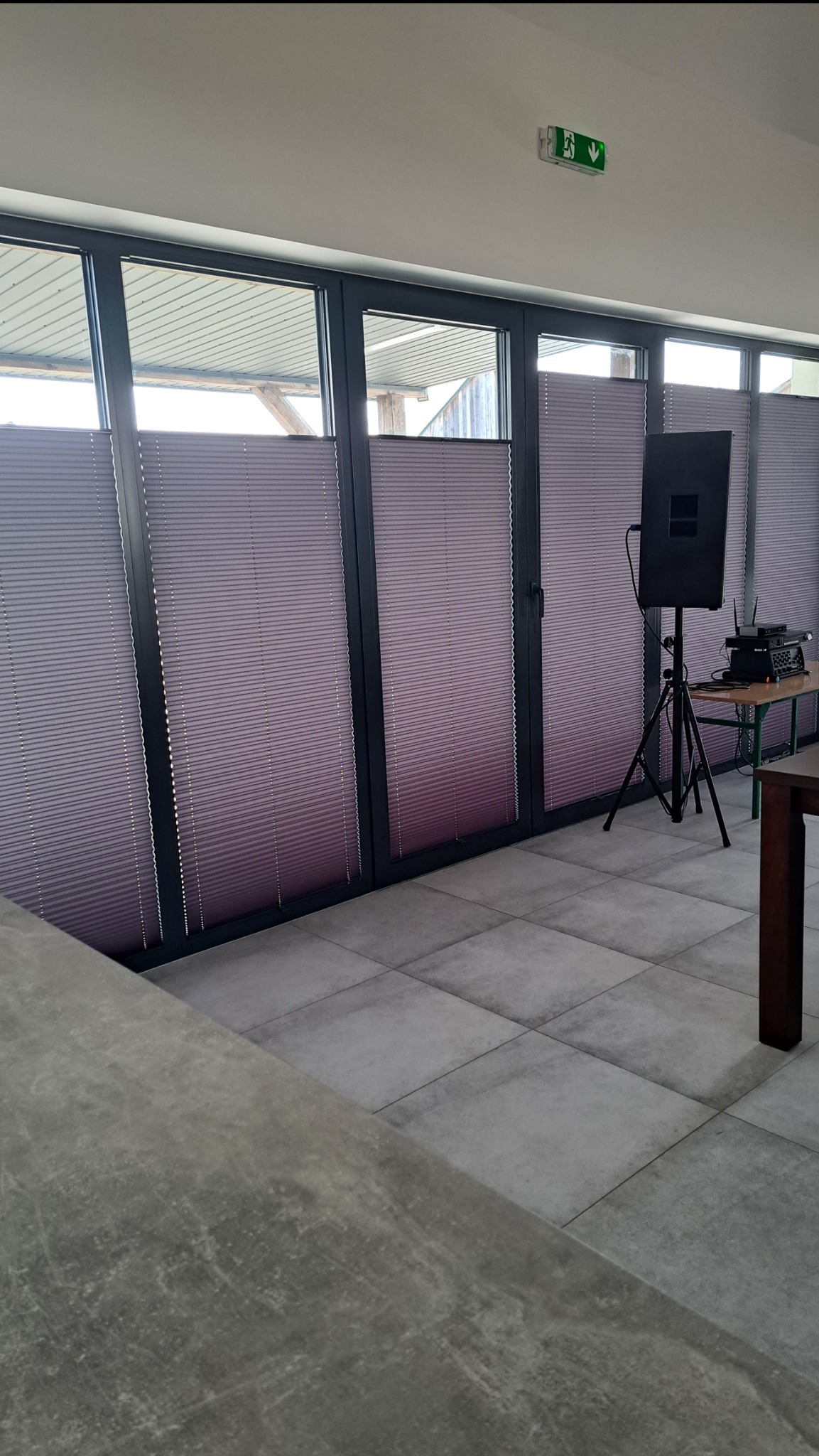 Fioletowe plisy okienne w szarej ramie, widoczne na tle jasnego pomieszczenia z szarą podłogą. Na drugim planie sprzęt nagłośnieniowy.