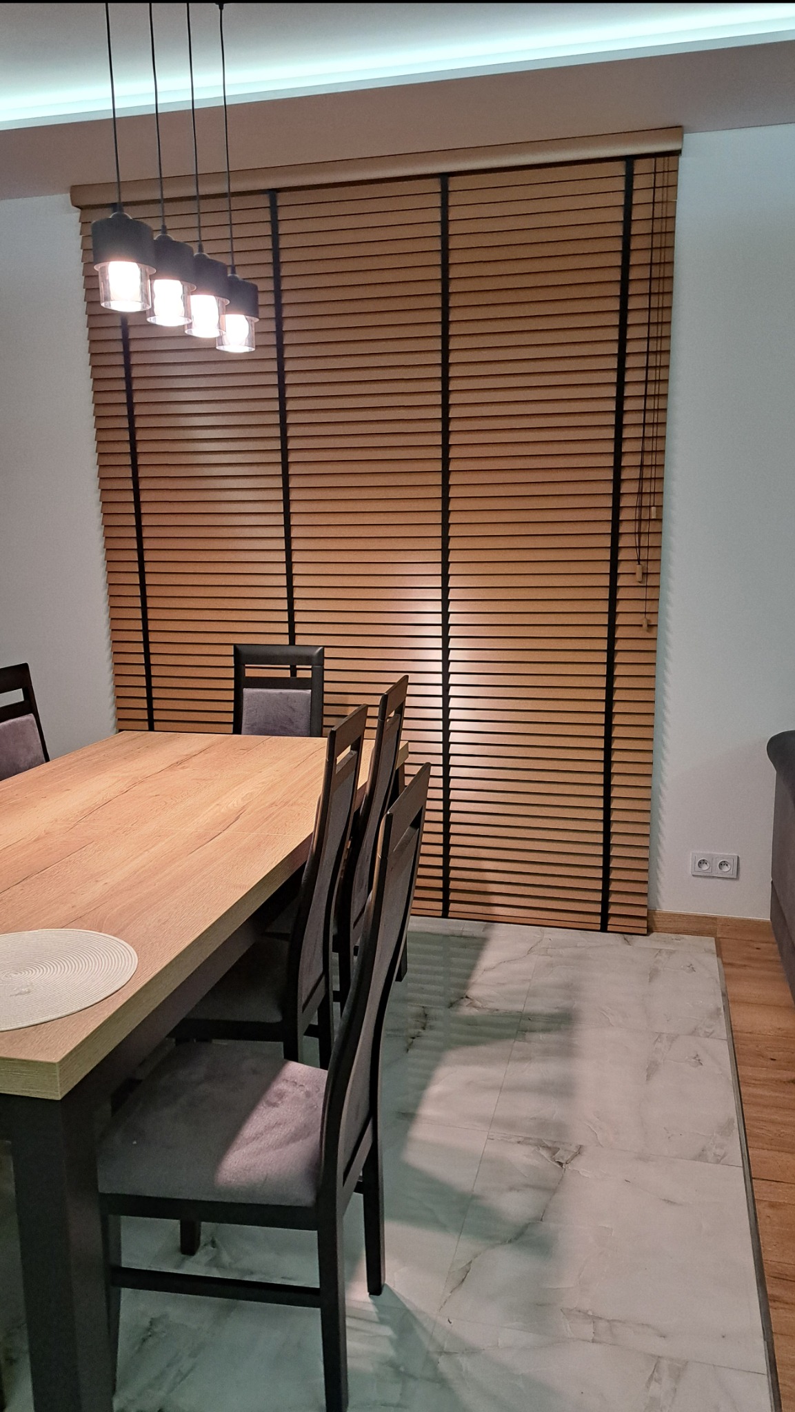 Drewniane żaluzje poziome w odcieniach brązu, zamontowane w oknie jadalni w Końskich. Widoczne czarne linki sterujące i fragment stołu z krzesłami.