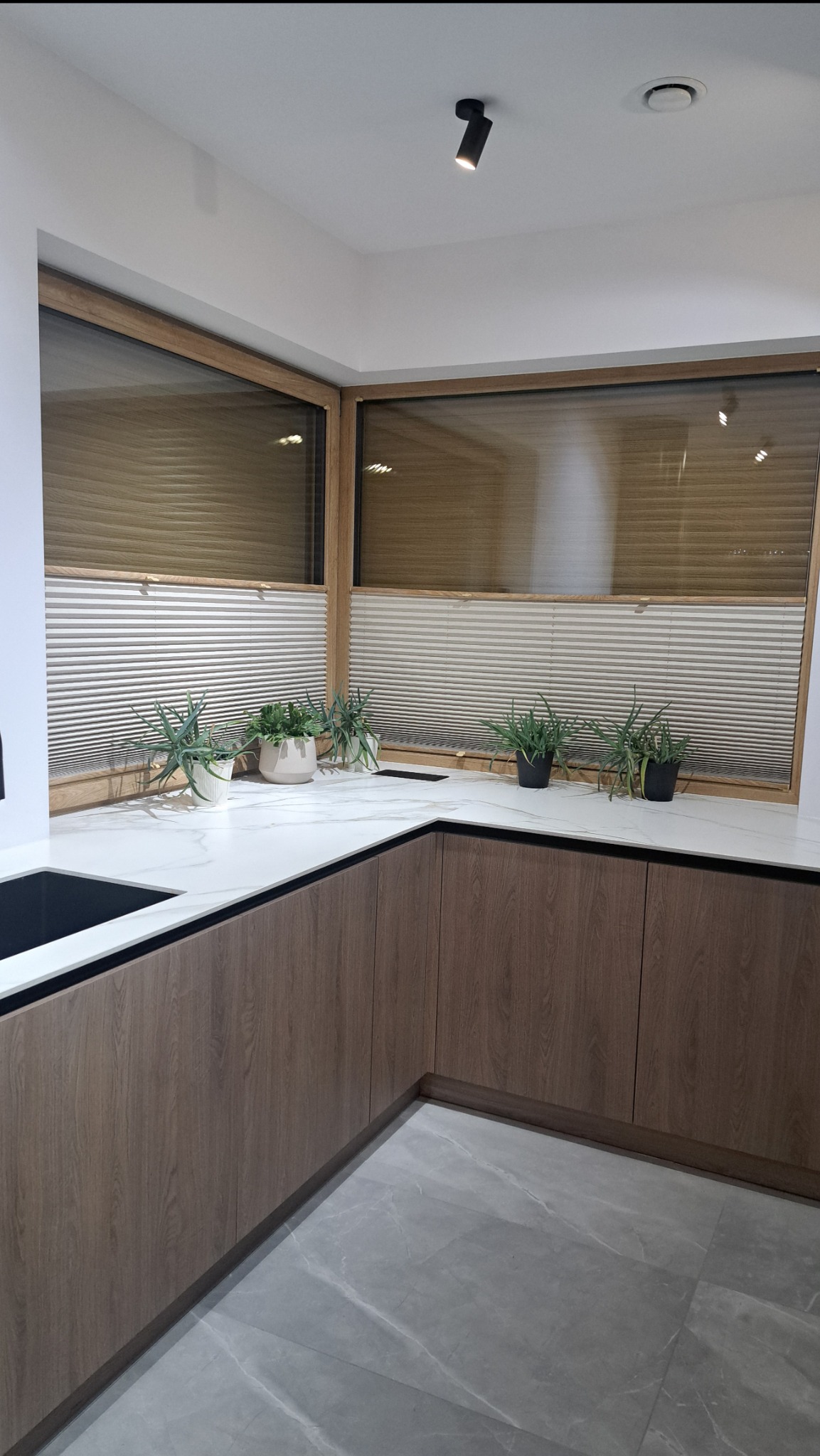 Jasne plisy okienne w drewnianych ramach, dekorowane zielonymi roślinami, na tle nowoczesnej kuchni z marmurowym blatem i drewnianymi szafkami. Minimalistyczny design.