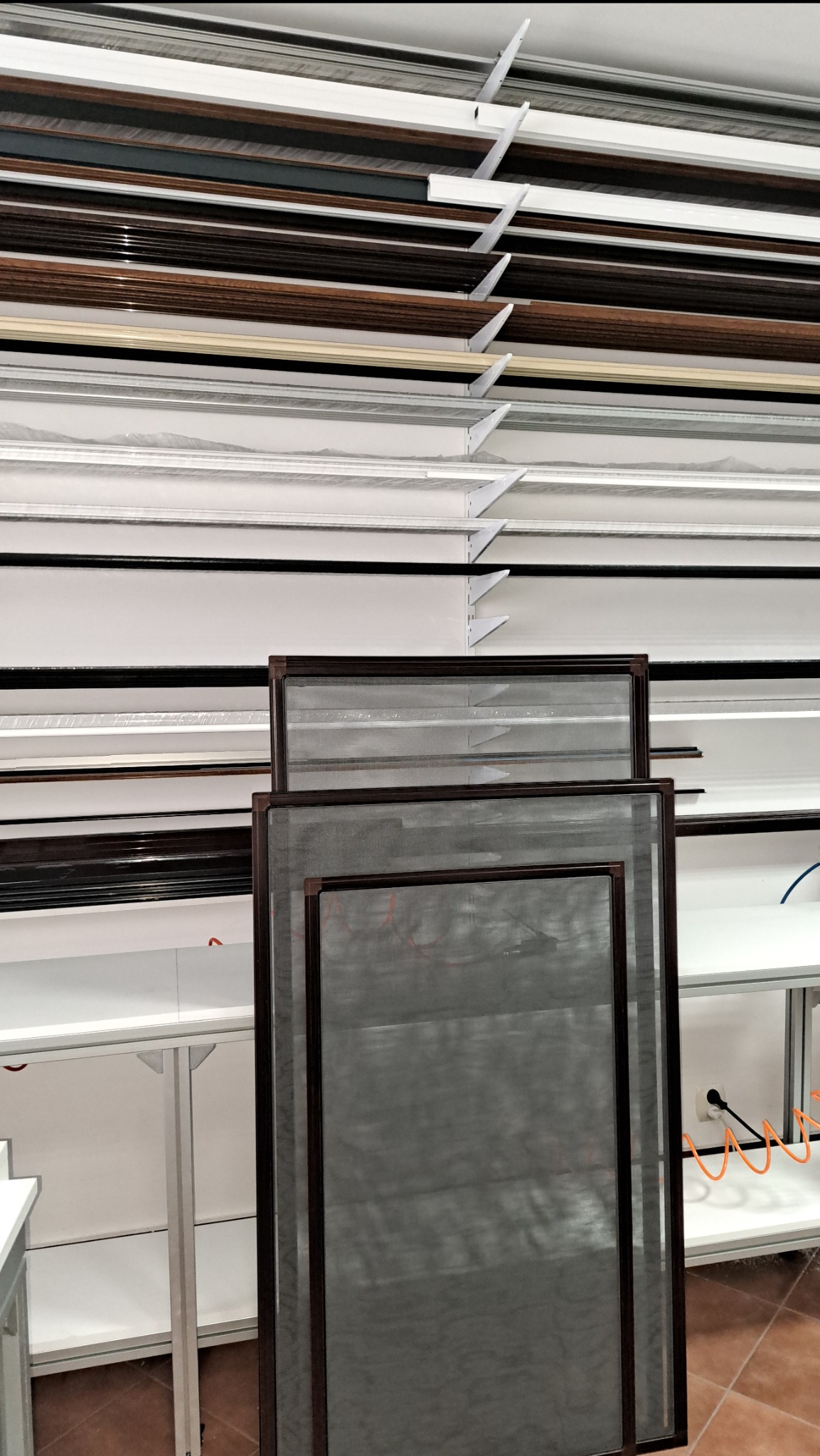 Dwie brązowe moskitiery na tle regału z ramami okiennymi o różnych kolorach. Widok z dołu, ukazujący różnorodność wykończeń.