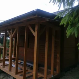 Drewniana konstrukcja z zadaszeniem i rynnami, prawdopodobnie w trakcie budowy, przylegająca do budynku z brązową elewacją, widok z zewnątrz w pochmurny dzień.