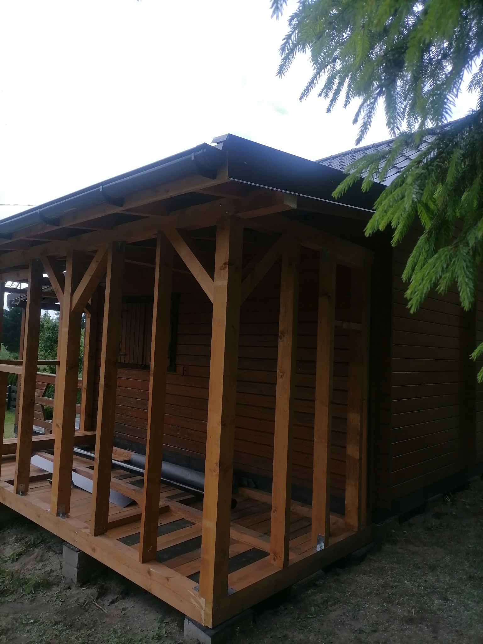 Drewniana konstrukcja z zadaszeniem i rynnami, prawdopodobnie w trakcie budowy, przylegająca do budynku z brązową elewacją, widok z zewnątrz w pochmurny dzień.