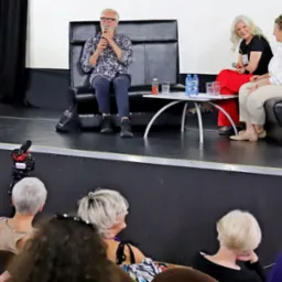 Konferencja prasowa na scenie: mężczyzna w kwiecistej koszuli przemawia do widowni, siedząc obok dwóch kobiet. Stolik z wodą i szklankami. Ujęcie z dołu, widoczna publiczność.