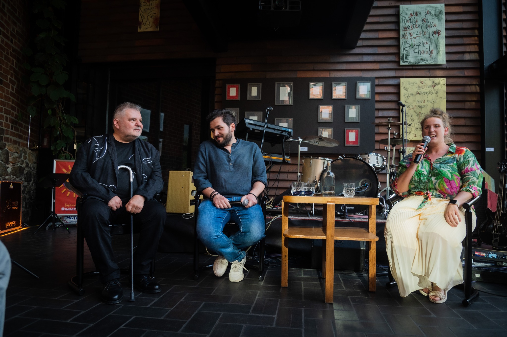 Panel dyskusyjny z trzema osobami siedzącymi na krzesłach na scenie z instrumentami muzycznymi w tle. Kobieta w kwiecistej koszuli trzyma mikrofon.