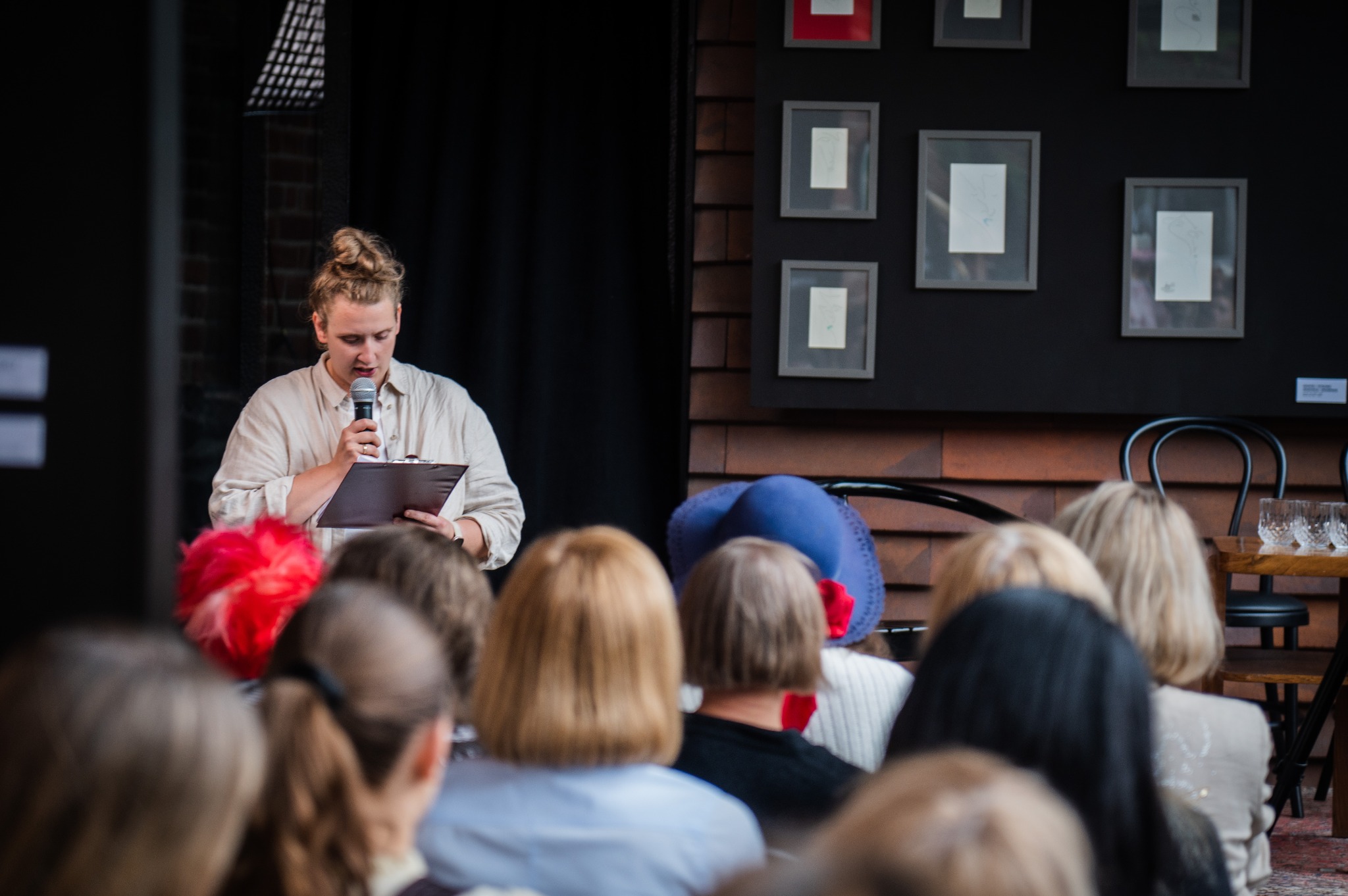 Prelegentka z mikrofonem na tle czarnej ściany z grafikami i publiczności. Kobieta w jasnej koszuli czyta z kartki. W tle rzędy słuchaczy.