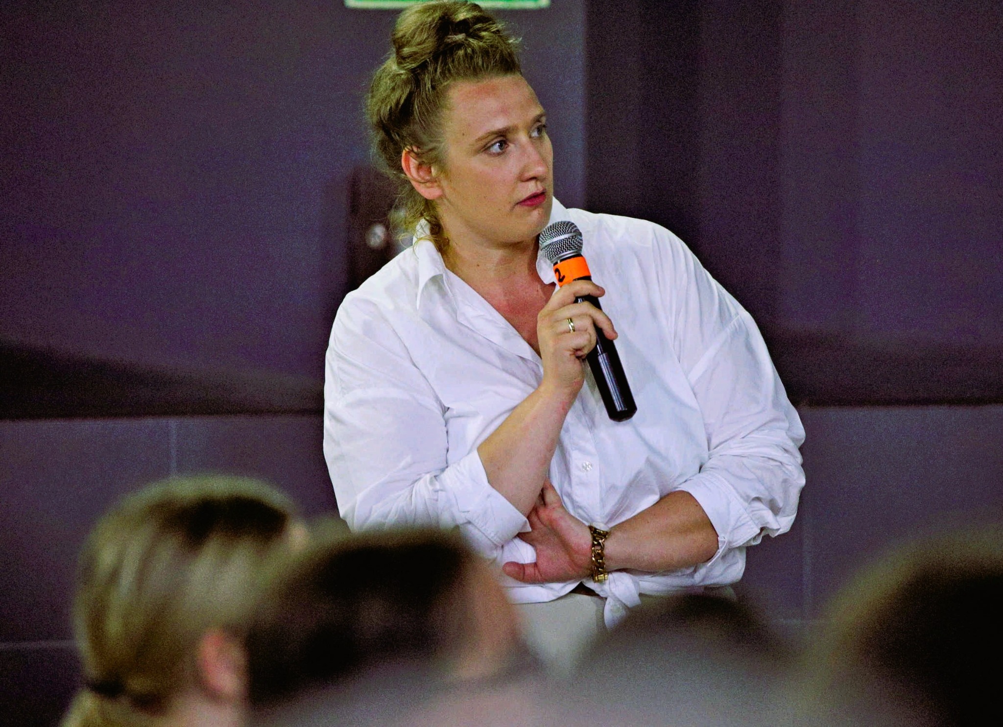 Kobieta z mikrofonem w białej koszuli, skupiona na mówieniu podczas prezentacji. W tle ciemna ściana i fragmenty widowni.