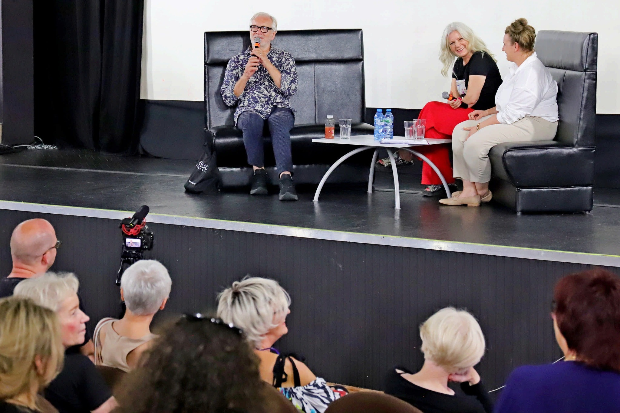 Konferencja prasowa na scenie: mężczyzna w kwiecistej koszuli przemawia do widowni, siedząc obok dwóch kobiet. Stolik z wodą i szklankami. Ujęcie z dołu, widoczna publiczność.