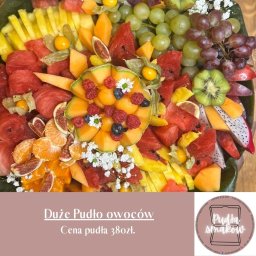 PUDŁA SMAKÓW - Monika Bijowska P.P.H.U 'MAJ-PLAST' - Bogata kompozycja owocowa: arbuz, melon, kiwi, figi, mandarynki, winogrona, miechunka, smoczy owoc i maliny z borówkami, udekorowana białymi kwiatami, tworząca apetyczną całość.