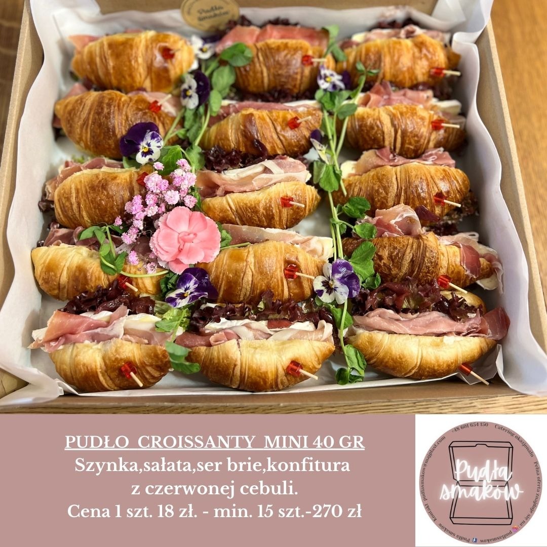 Dekoracyjne mini croissanty z szynką, serem brie i czerwoną cebulą, ułożone w pudełku, ozdobione kwiatami. Idealne na catering okolicznościowy.