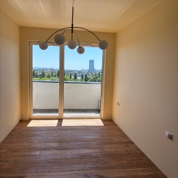 TK Usługi Budowlane - Jasne wnętrze pokoju z drewnianą podłogą, widokiem na miasto z balkonu i nowoczesną lampą sufitową. Ściany pomalowane na kremowy kolor.