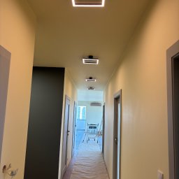 TK Usługi Budowlane - Pomalowany korytarz z nowym oświetleniem LED. Ściany w odcieniach beżu, podłoga zabezpieczona folią malarską. Widoczny wózek malarski w oddali.
