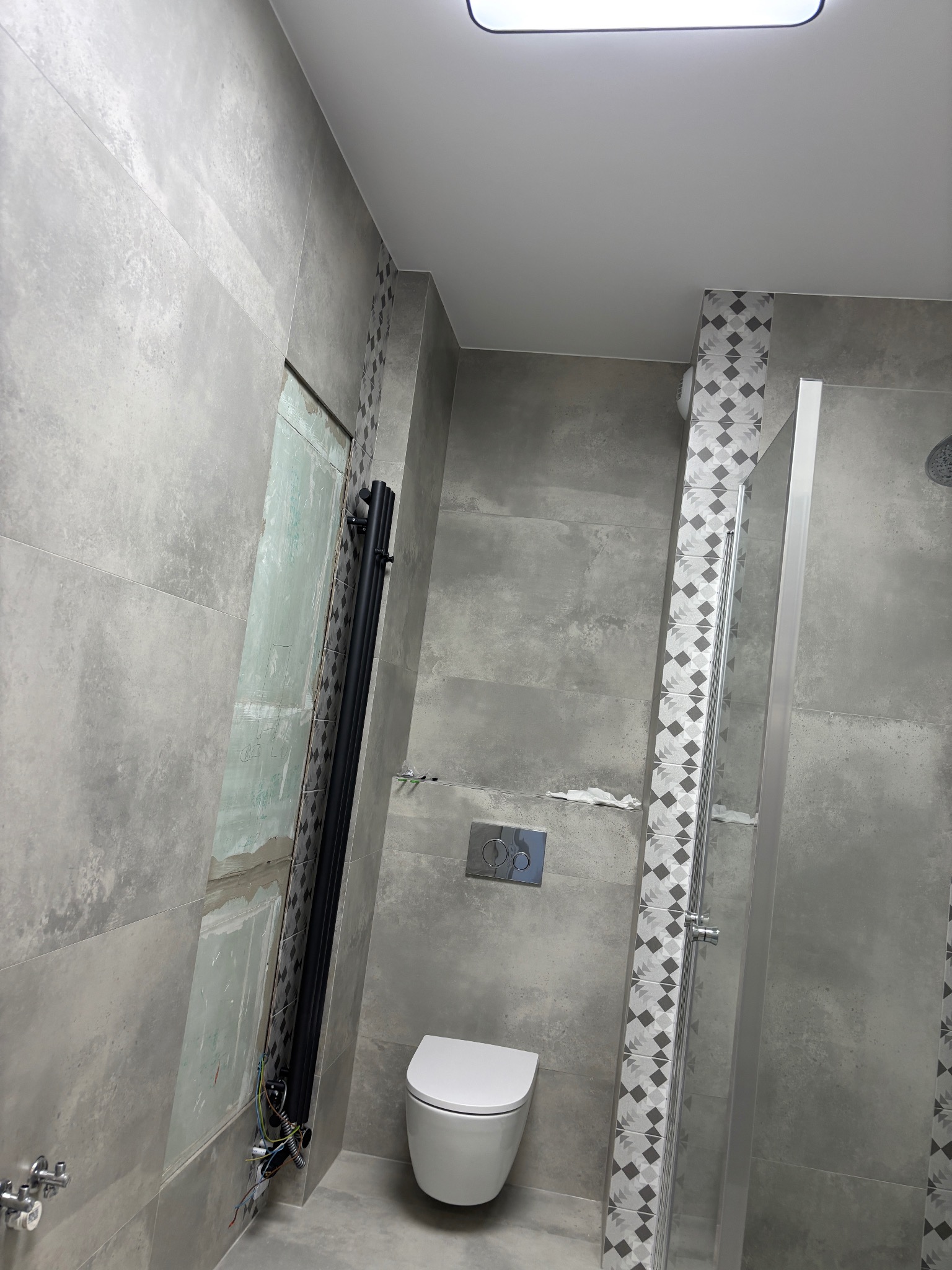 Szara łazienka w trakcie wykańczania: widoczna toaleta, kabina prysznicowa z dekoracyjną mozaiką, grzejnik drabinkowy i fragment ściany bez płytek.