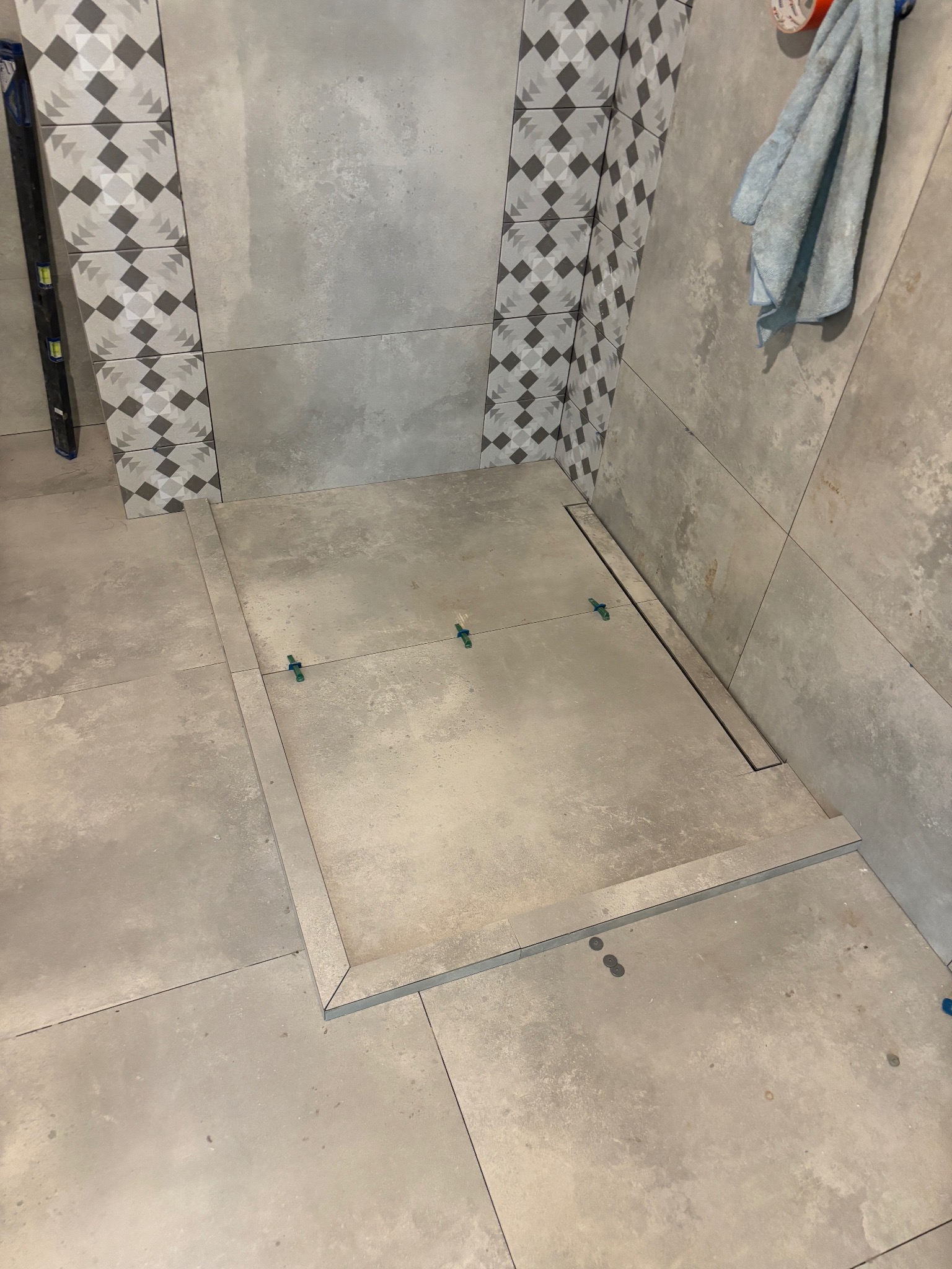 Ułożone szare płytki w łazience z odpływem liniowym w strefie prysznicowej. Widoczne krzyżyki poziomujące. Na ścianie dekoracyjne płytki w romby.