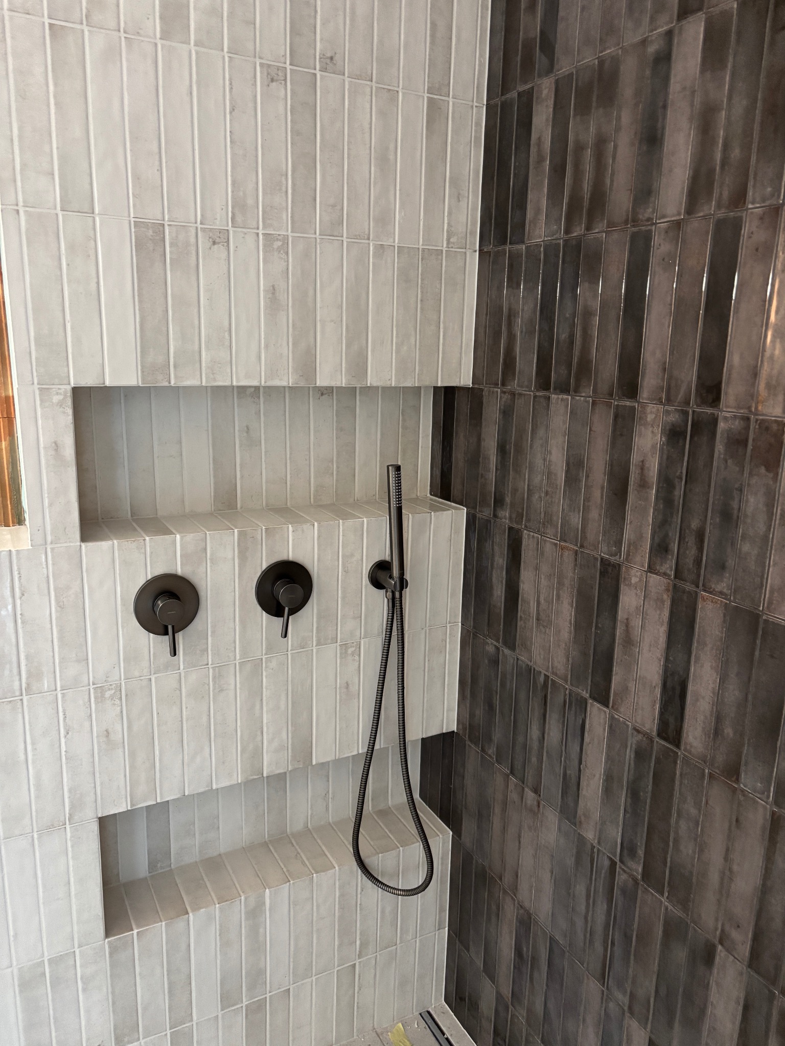 Nowoczesna kabina prysznicowa z jasnymi i ciemnymi, pionowo ułożonymi płytkami. Wnęki w ścianie wyłożone jasnymi płytkami. Czarna armatura.