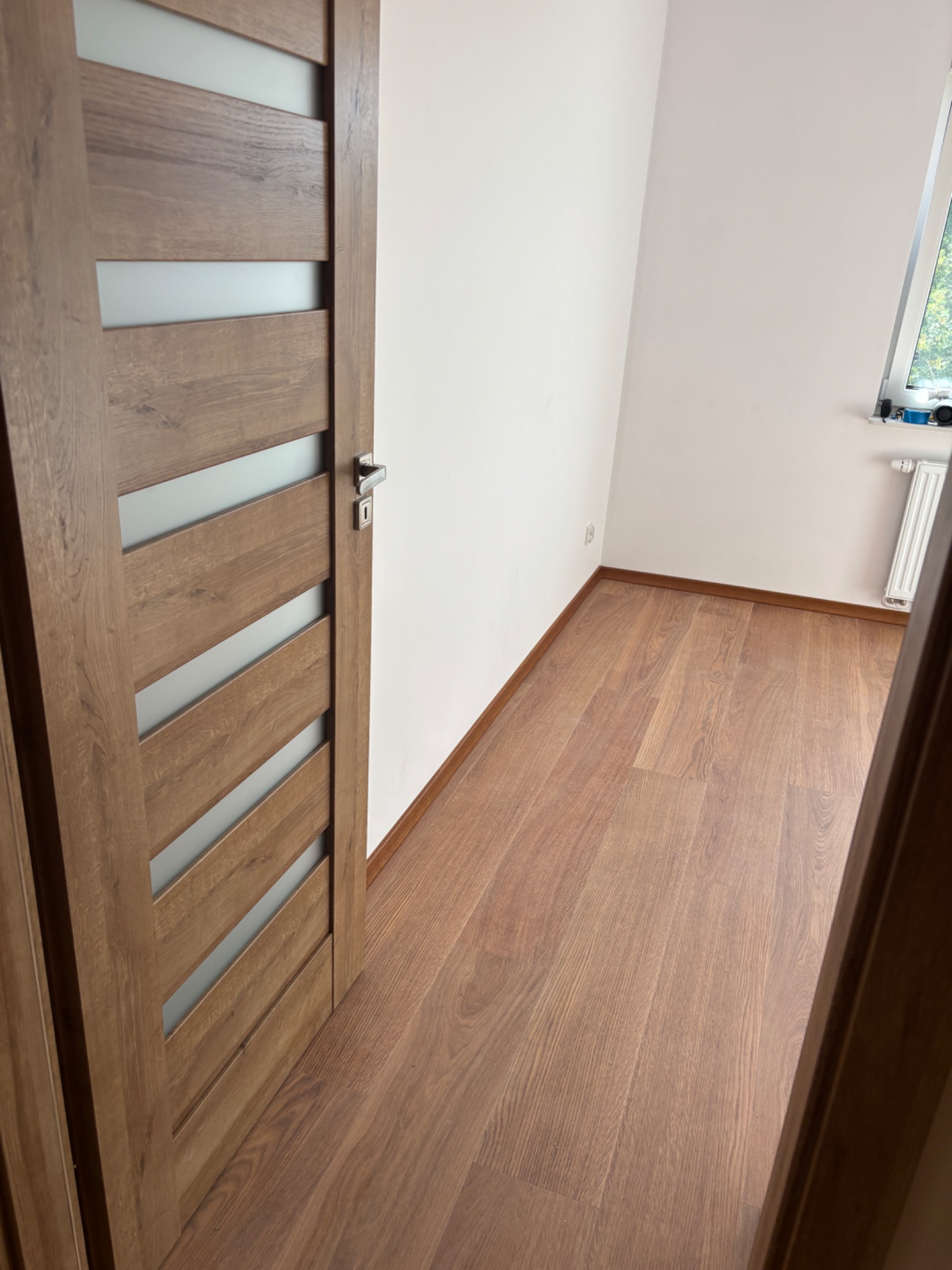 Otwarta, drewniana, wewnętrzna  drzwi z przeszkleniami, ukazująca wykończone wnętrze z drewnianą podłogą i białymi ścianami. Widoczny fragment okna i grzejnika.