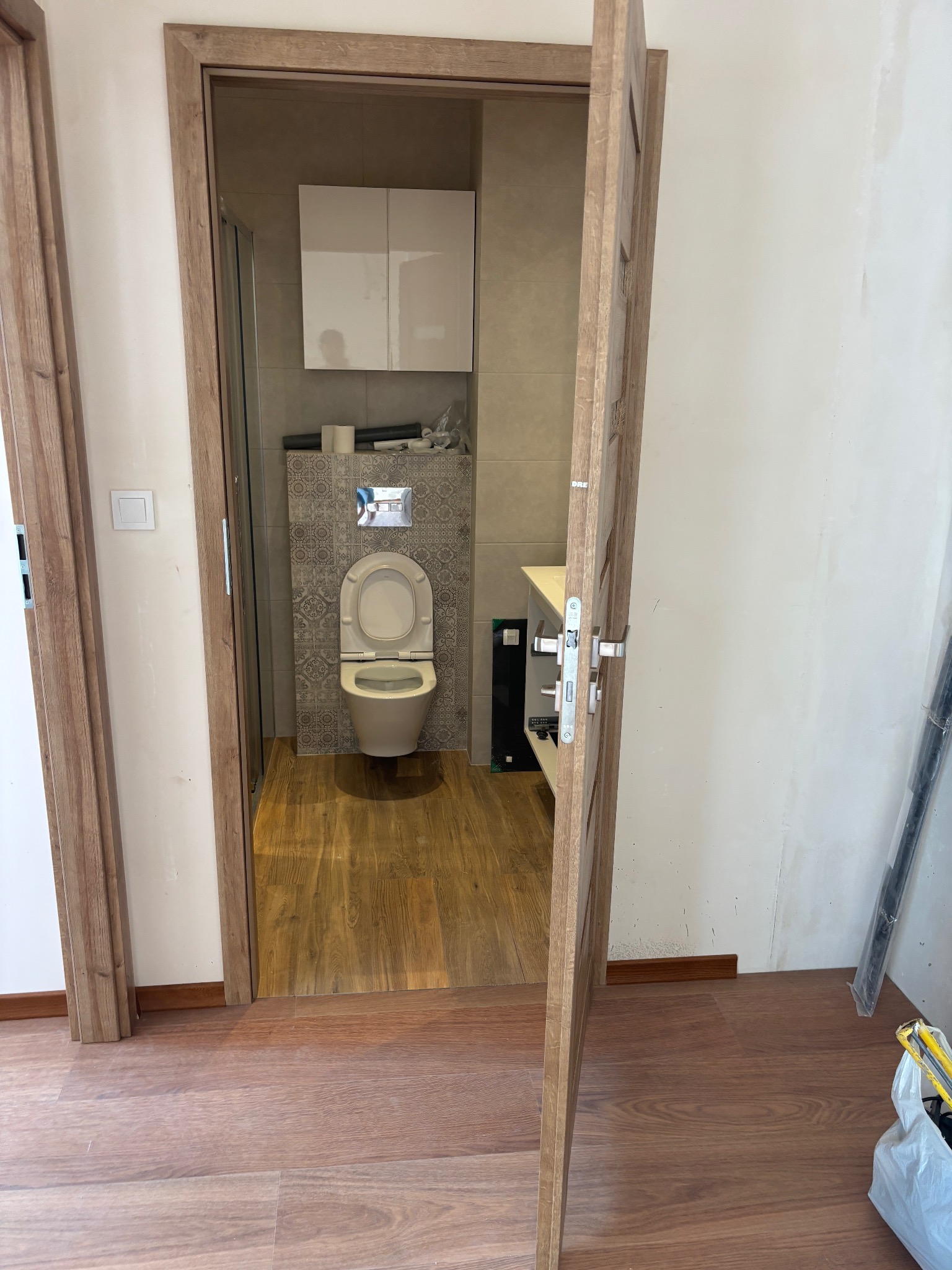 Wykończona łazienka z drewnopodobną podłogą, widoczna przez otwarte drzwi z drewnianą ościeżnicą. Na ścianie nad toaletą dekoracyjne płytki i szafka.