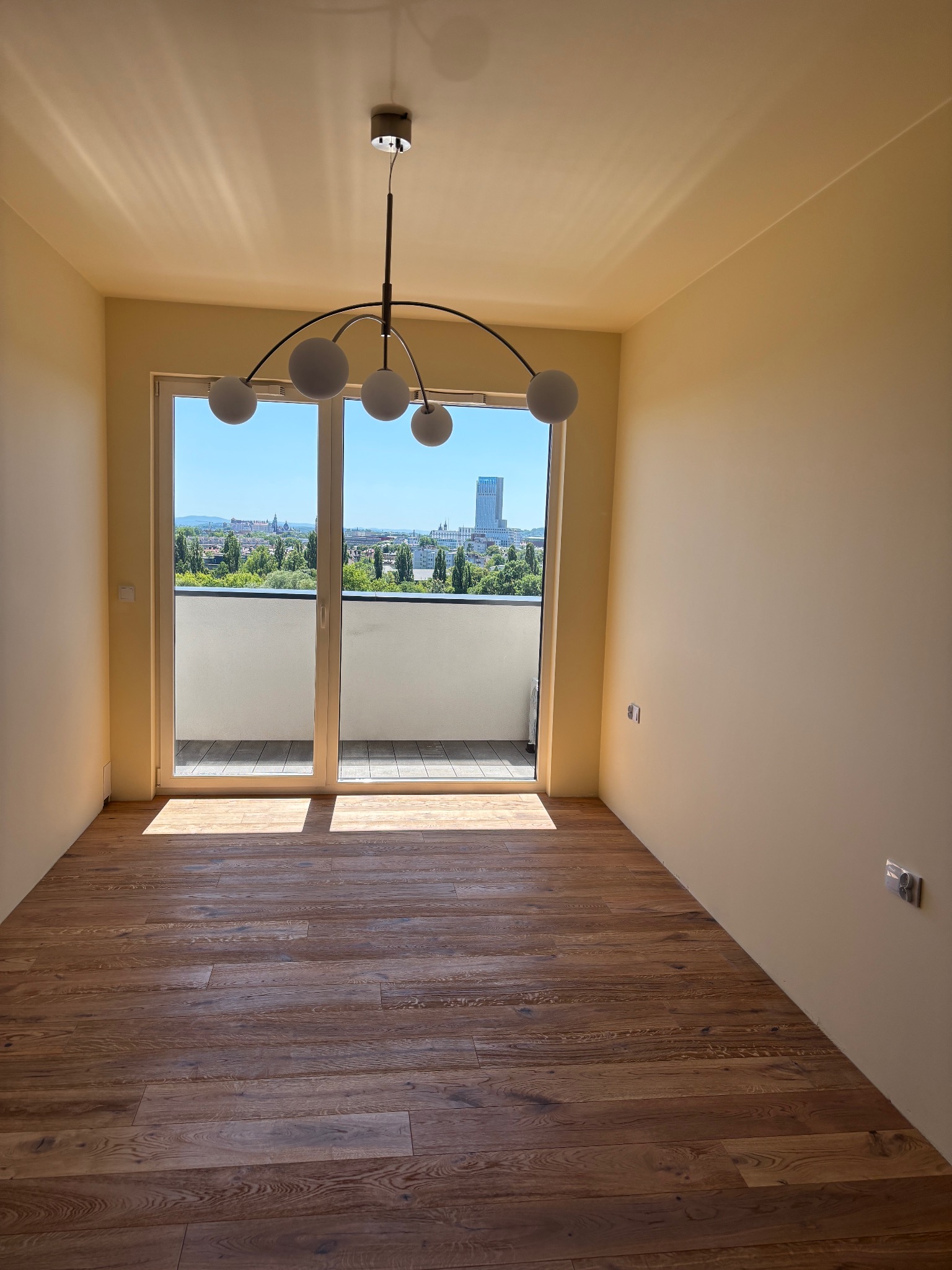Jasne wnętrze pokoju z drewnianą podłogą, widokiem na miasto z balkonu i nowoczesną lampą sufitową. Ściany pomalowane na kremowy kolor.