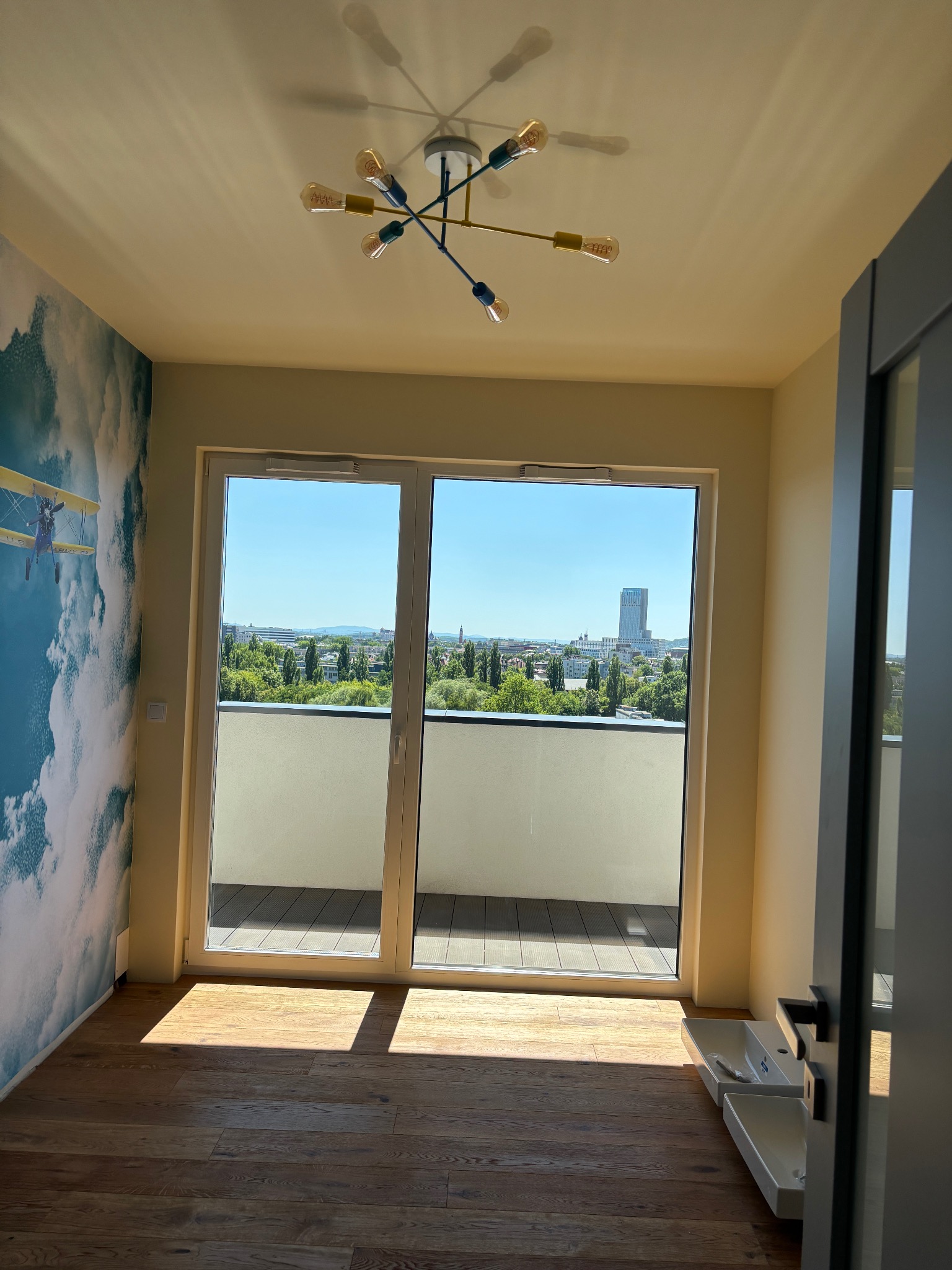 Pokój z drewnianą podłogą i tapetą w chmury z samolotem, widok na balkon i miasto. Na suficie nowoczesna lampa. Częściowo widoczna umywalka.