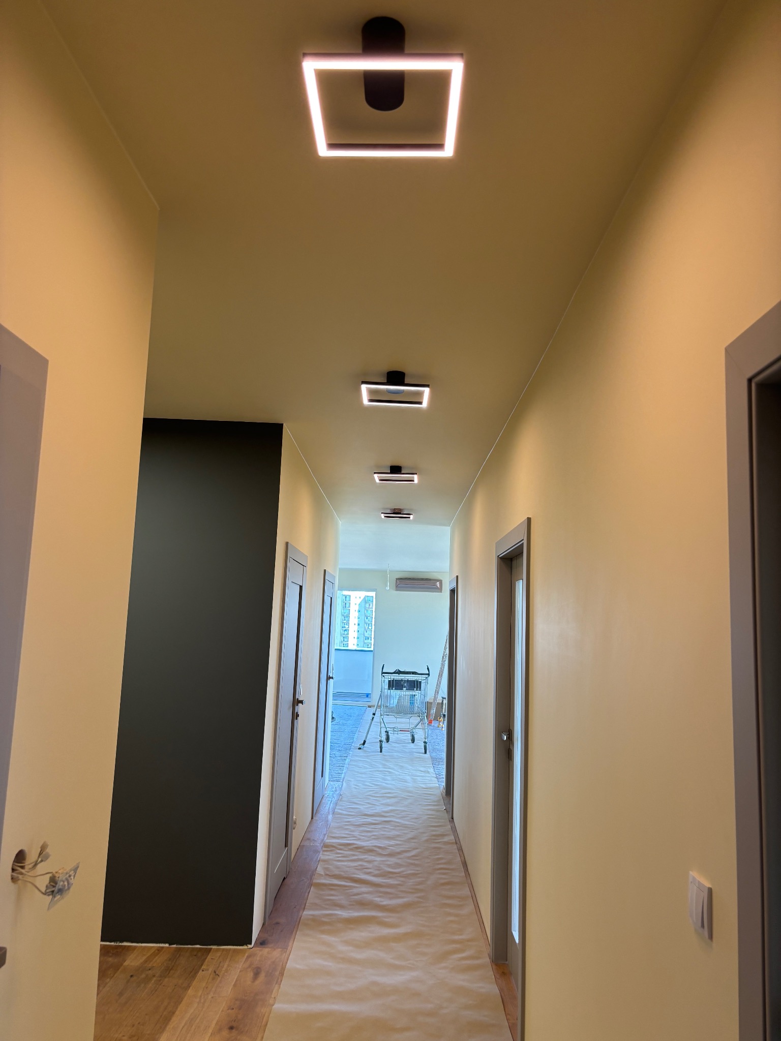 Pomalowany korytarz z nowym oświetleniem LED. Ściany w odcieniach beżu, podłoga zabezpieczona folią malarską. Widoczny wózek malarski w oddali.