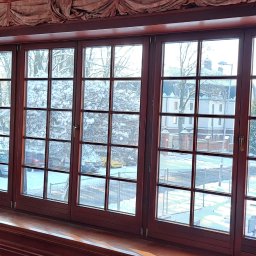 Sylwia Hahn Perfekt Home - Duże, drewniane okna szprosowe z widokiem na zimowy krajobraz. Widoczne śnieżne drzewa, budynki i samochody. Okno z ozdobną zasłoną i parapetem.