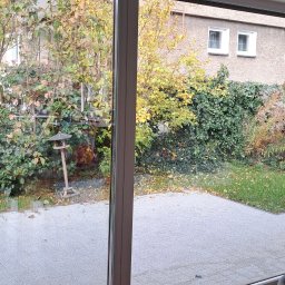 Sylwia Hahn Perfekt Home - Przesuwne okno tarasowe, widok na jesienny ogród z karmnikiem dla ptaków. Czysta szyba, szara kostka brukowa na tarasie.