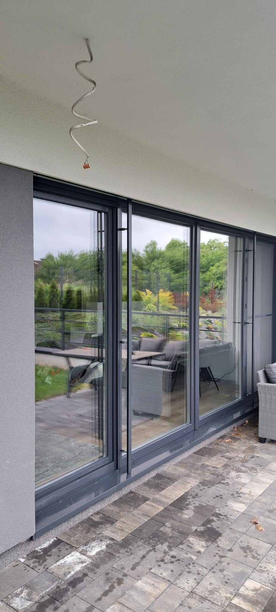 Panoramiczne okno tarasowe z szarymi ramami odbija zielony ogród. Widoczne ślady po deszczu na kostce brukowej i niedokończona instalacja na suficie.