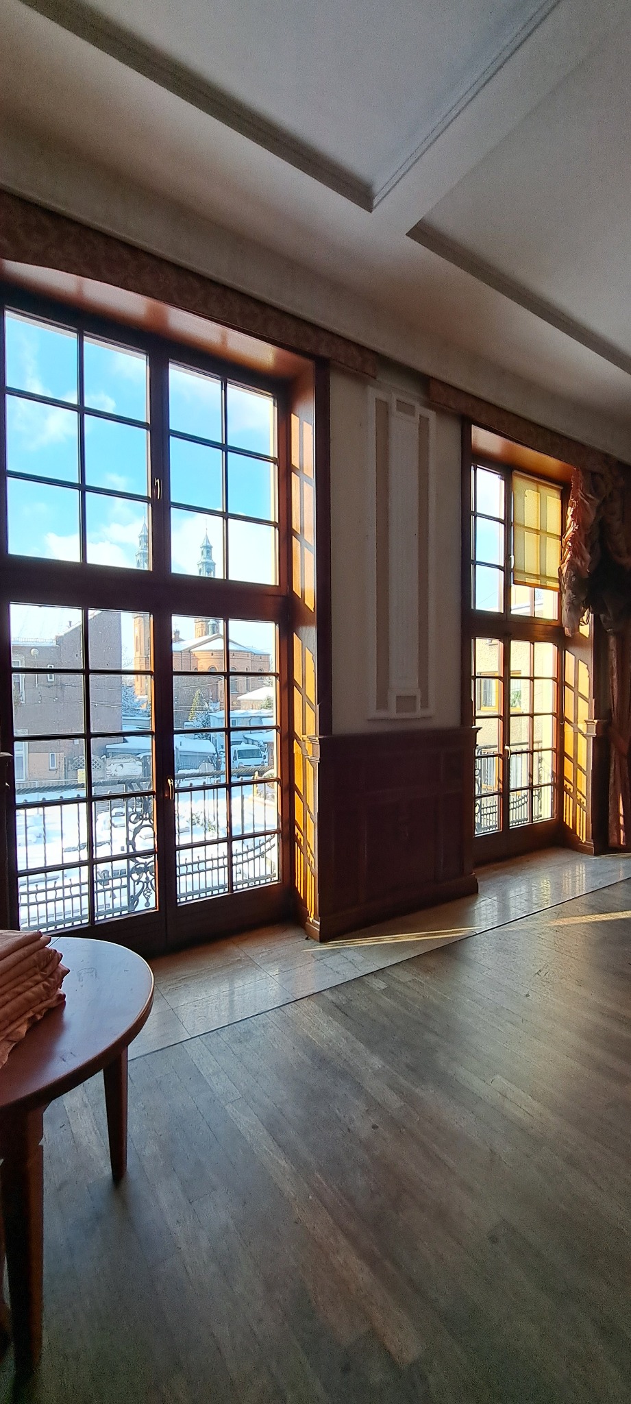 Sala z dużymi oknami w drewnianych ramach, przez które widać budynki. Na podłodze leżą promienie słoneczne. Widoczny fragment stołu z tkaniną.