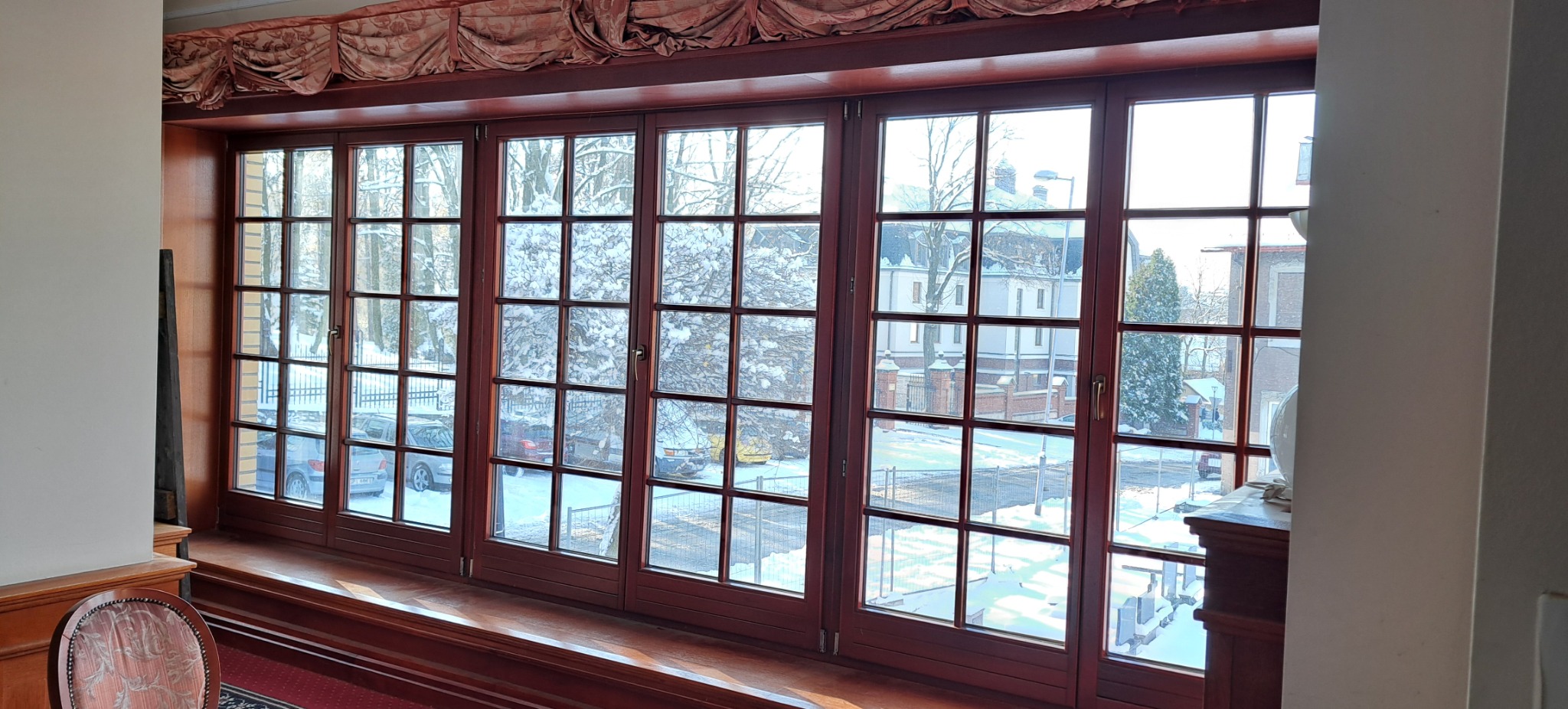 Duże, drewniane okna szprosowe z widokiem na zimowy krajobraz. Widoczne śnieżne drzewa, budynki i samochody. Okno z ozdobną zasłoną i parapetem.