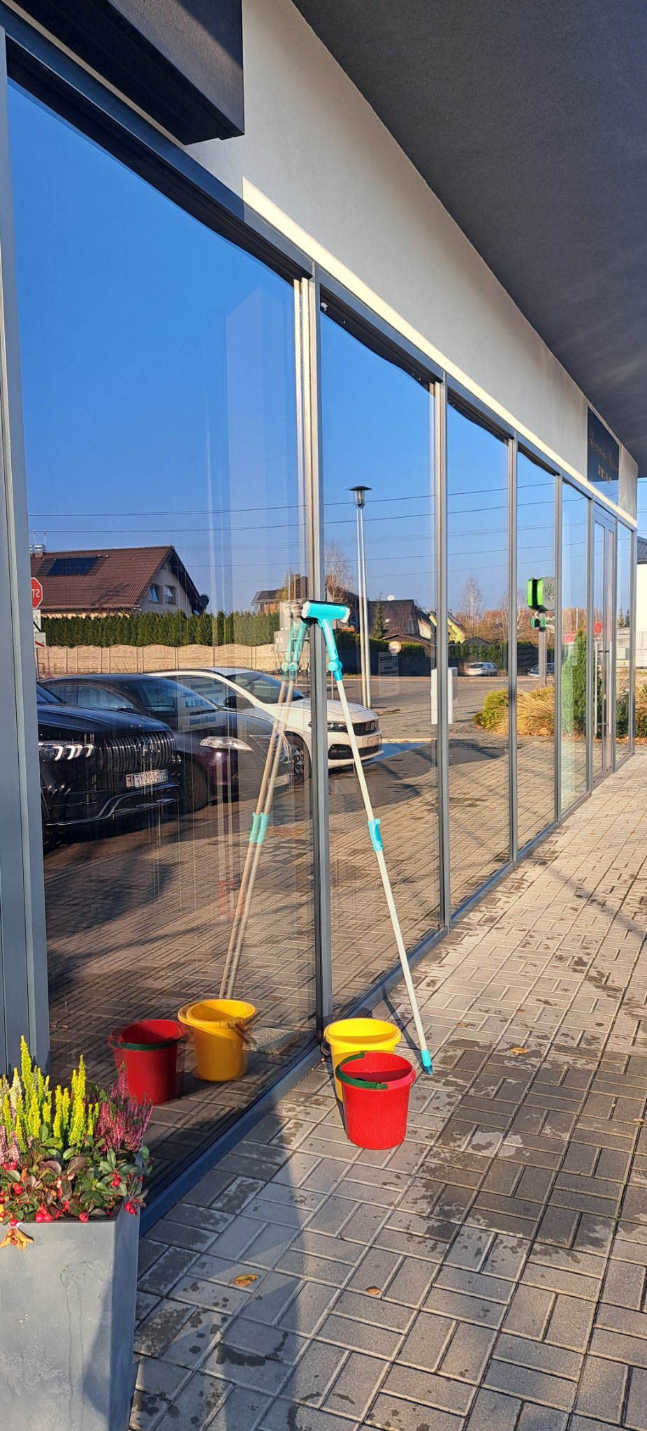 Mycie okien w lokalu usługowym: widoczne wiaderka, myjka i odbicie otoczenia w czystych szybach. Słoneczny dzień, kostka brukowa.