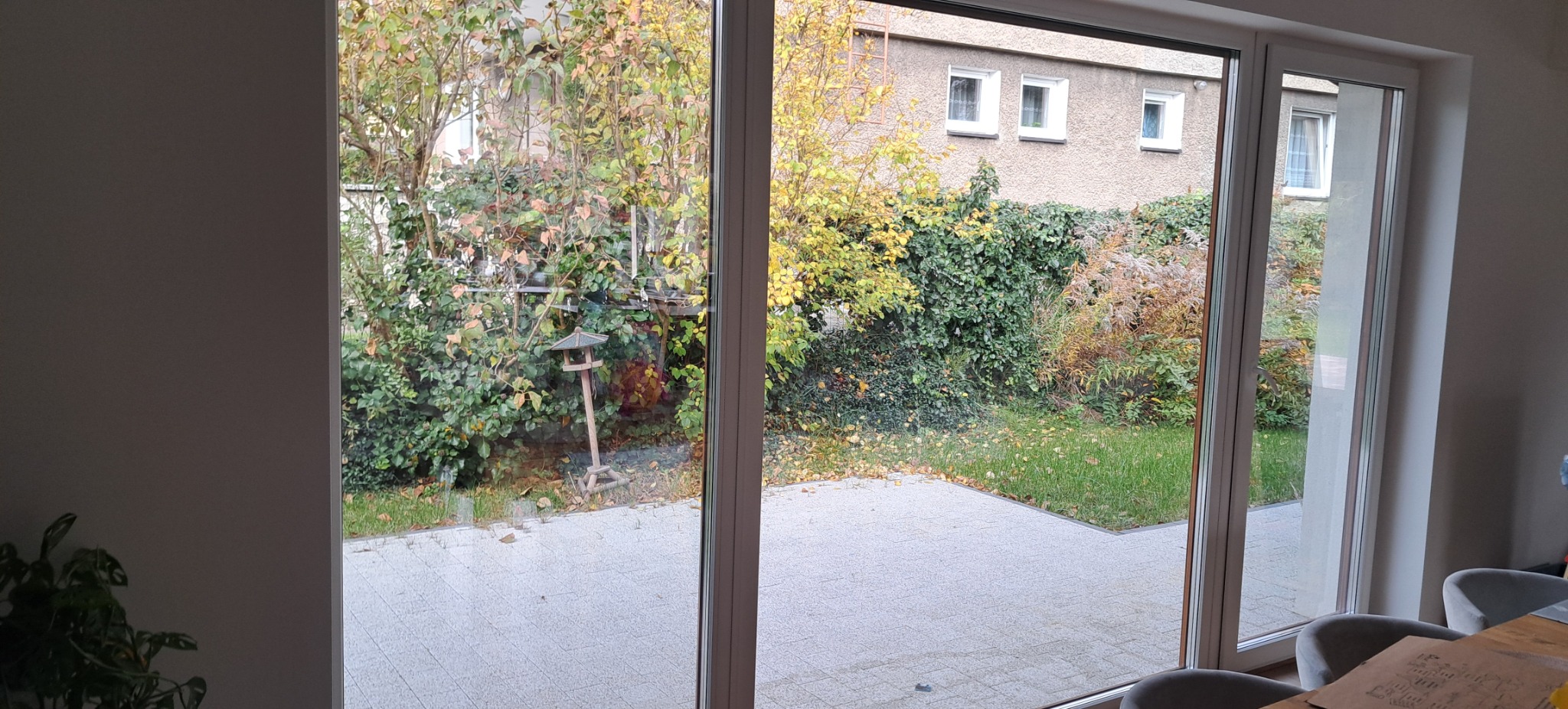 Przesuwne okno tarasowe, widok na jesienny ogród z karmnikiem dla ptaków. Czysta szyba, szara kostka brukowa na tarasie.