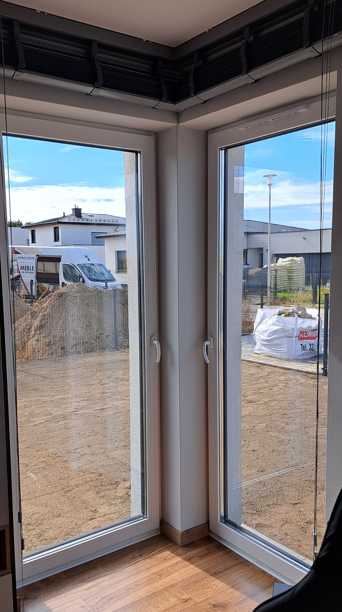 Kątowe okno z widokiem na plac budowy. Na zewnątrz biały bus z napisem 'Nowoczesne Meble na Wymiar' oraz worki z materiałami budowlanymi.