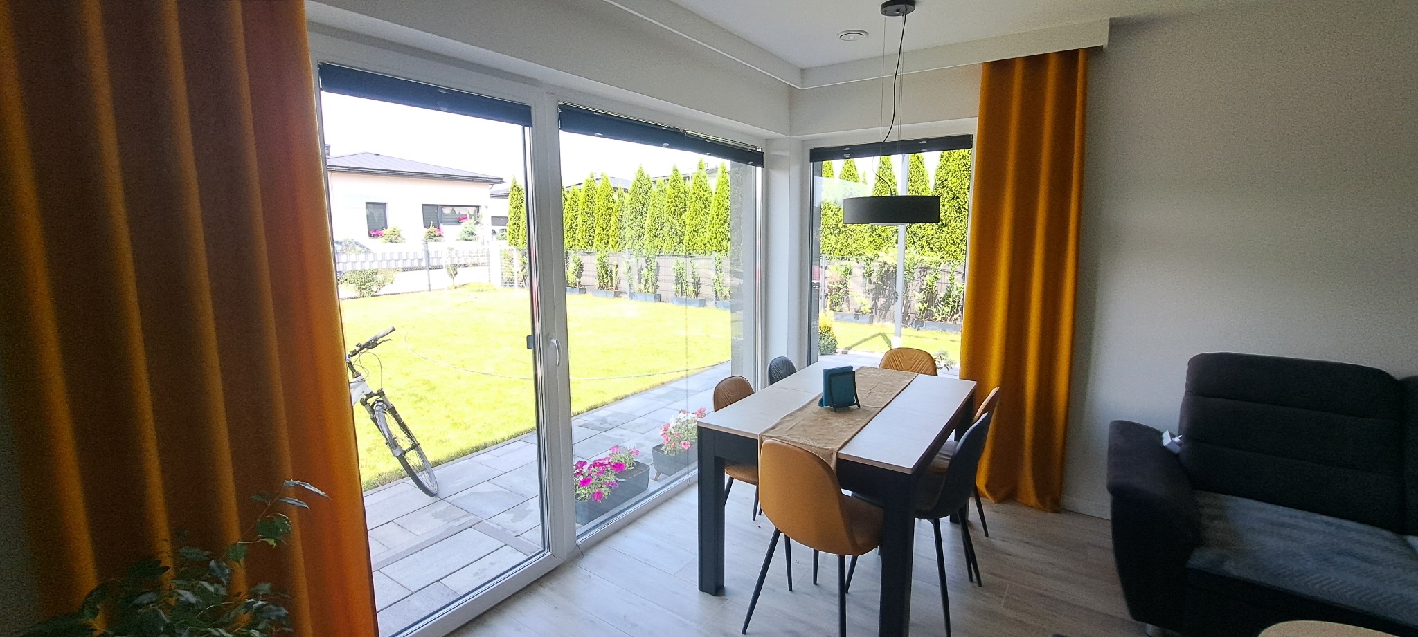 Jasne wnętrze jadalni z dużym oknem na ogród, przez które widać rower oparty o ścianę. Żółte zasłony po bokach okna dodają ciepła wnętrzu.