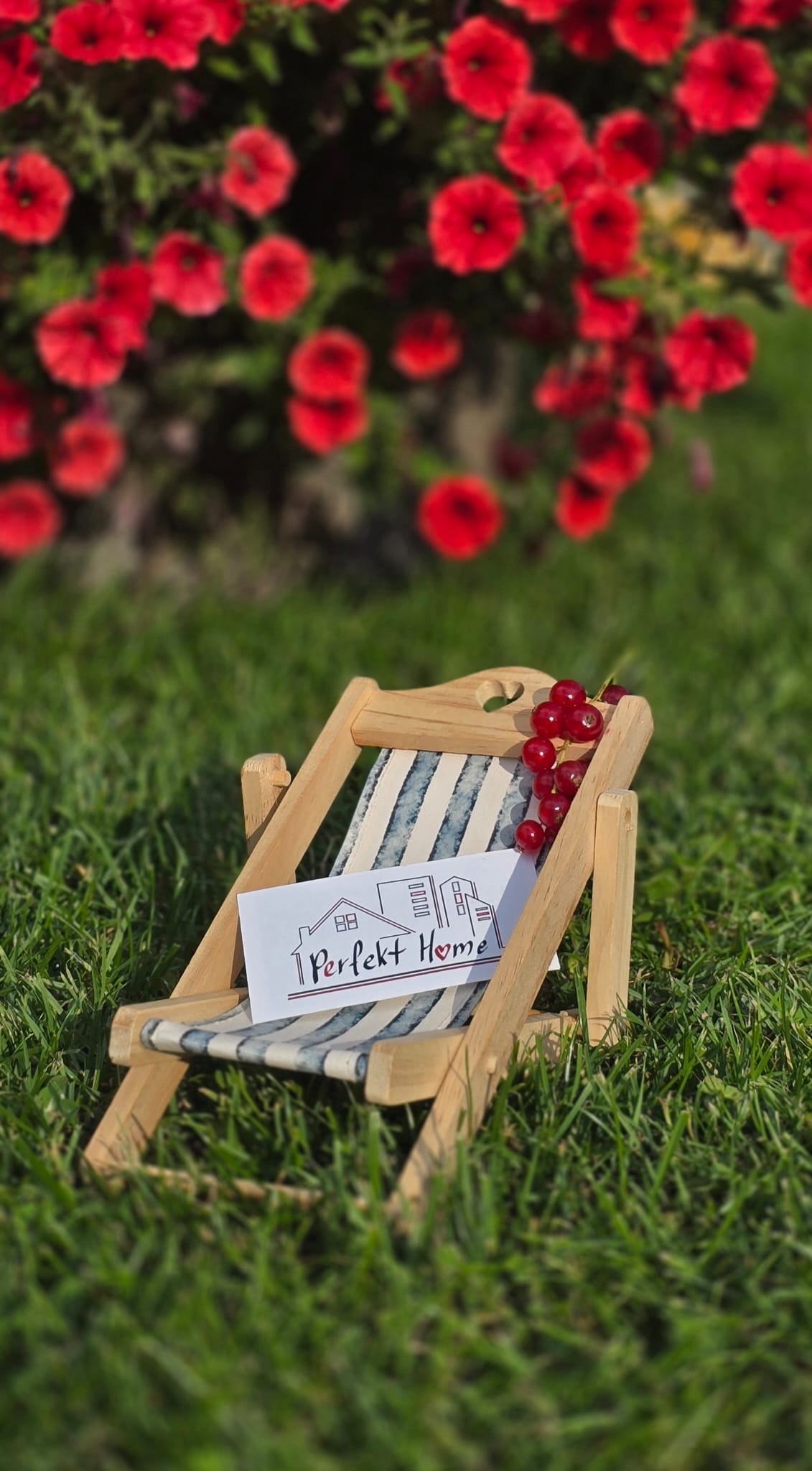 Miniaturowy leżak z wizytówką 'Perfekt Home' na trawie, czerwone kwiaty w tle. Nietypowe ujęcie z bliska, słoneczny dzień.