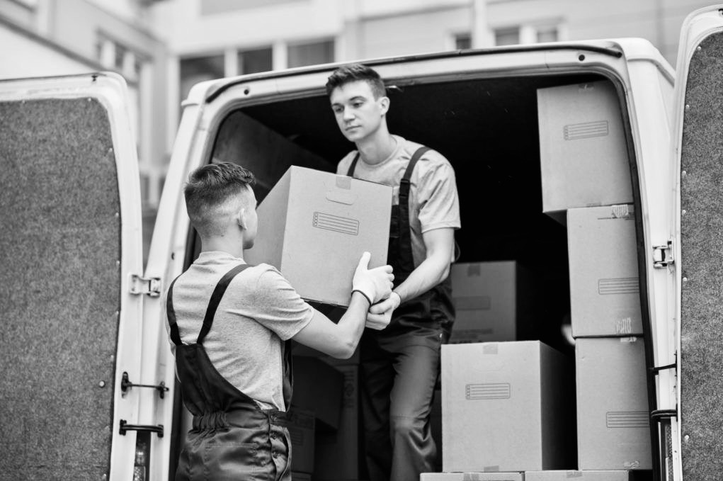Dwóch mężczyzn w kombinezonach przenosi kartonowe pudła do wnętrza furgonetki. Monochromatyczne ujęcie z otwartymi drzwiami pojazdu ukazuje etap załadunku.