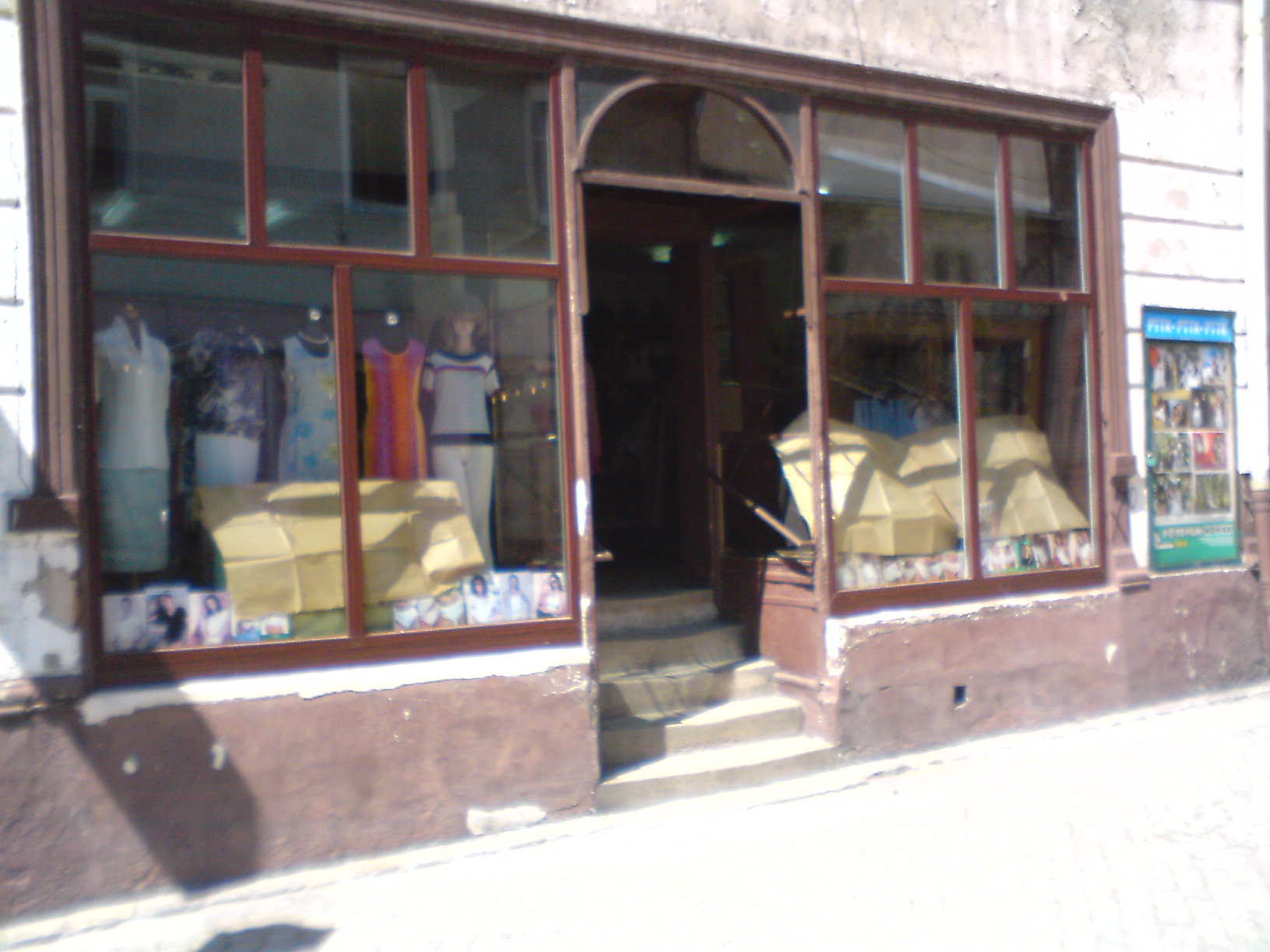 Witryna lokalu użytkowego w Połczynie-Zdroju z manekinami w ubraniach i piramidami z kartonów. Widoczne schody wejściowe i plakat reklamowy po prawej stronie.