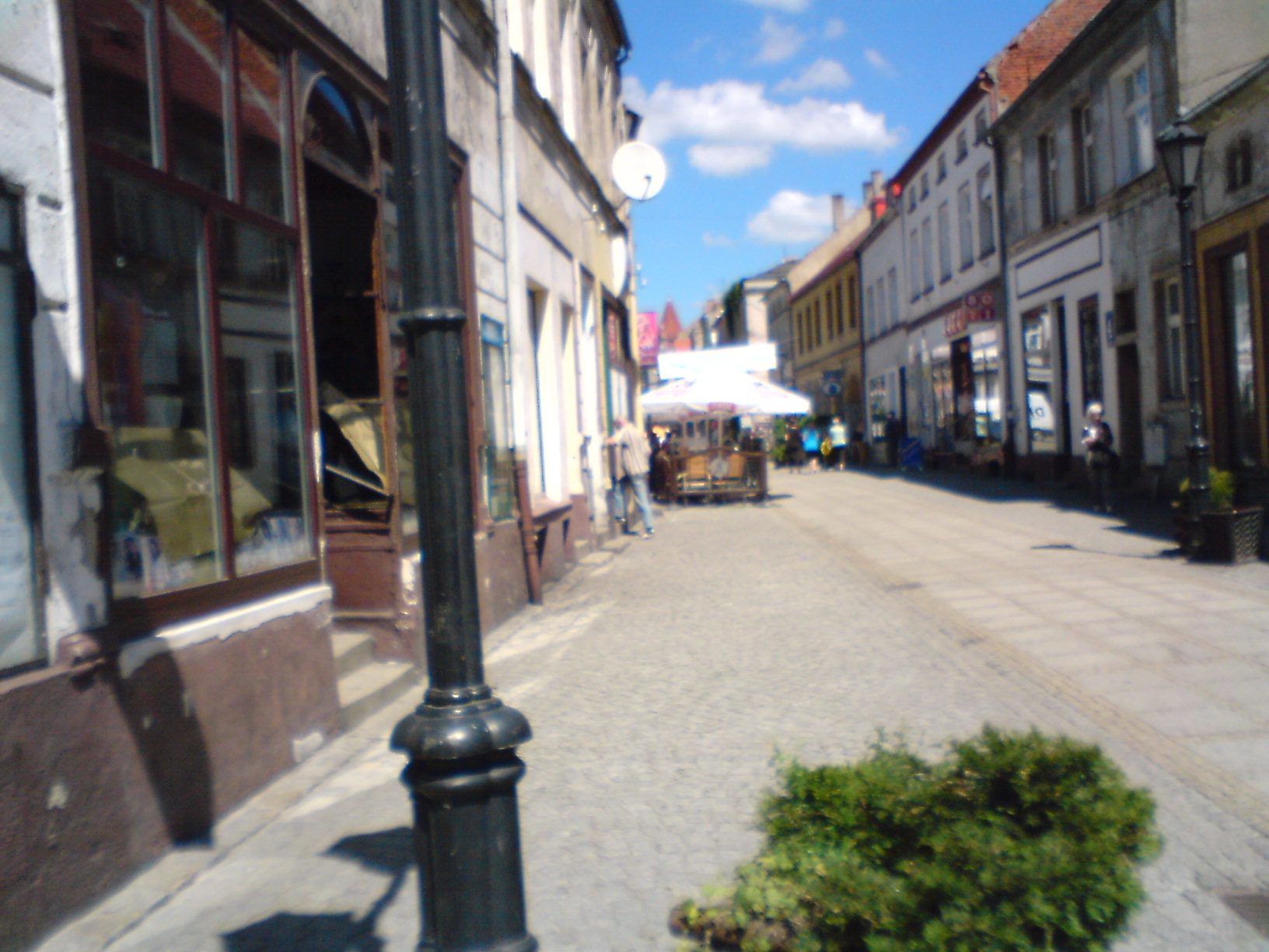 Ulica w małym miasteczku z widocznymi lokalami użytkowymi, jeden z nich ma otwarte drzwi i okna, w tle ogródek letni pod białym parasolem.