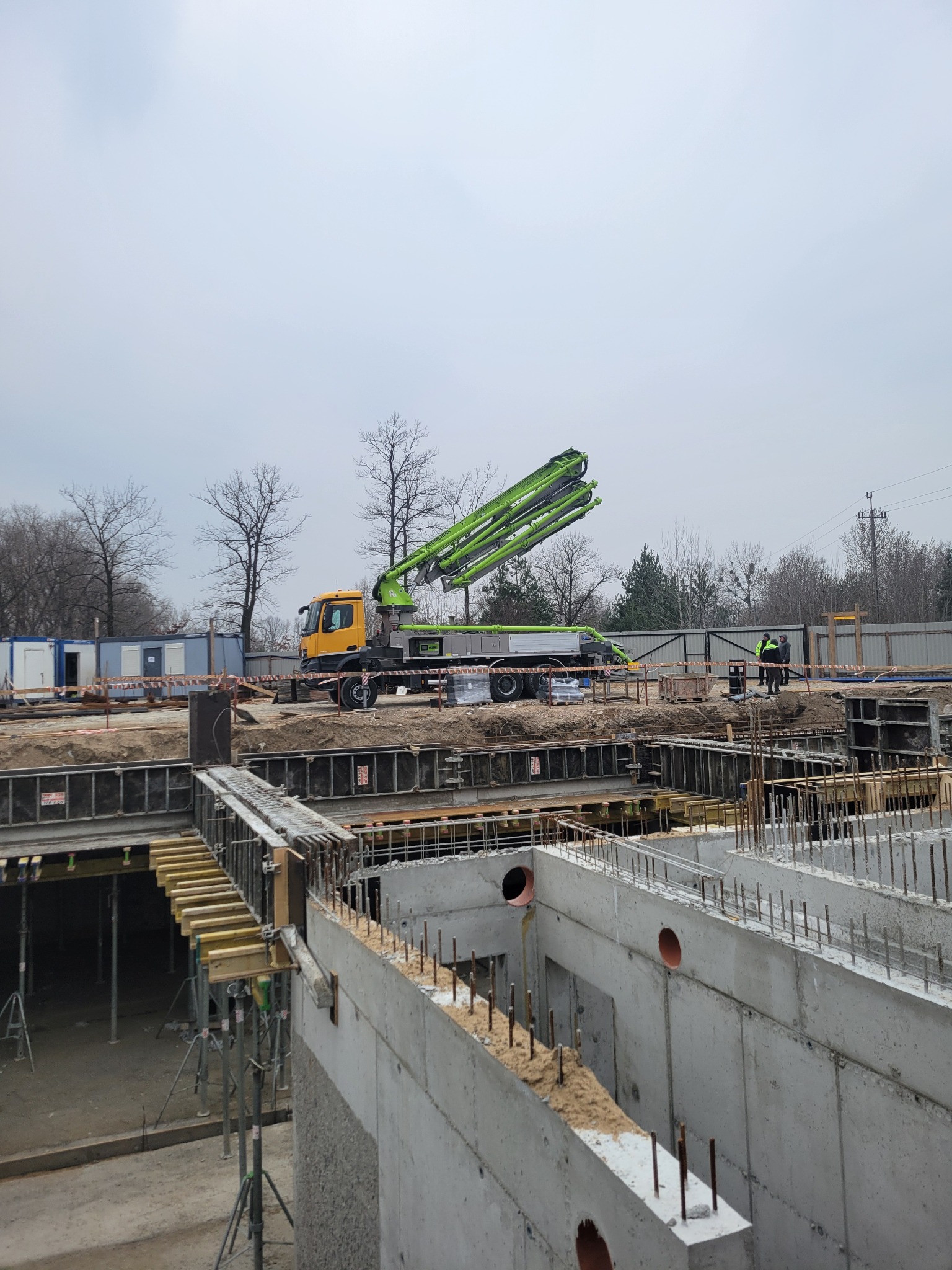 Wykop fundamentów z betonowymi ścianami szalunkowymi i zbrojeniem, na budowie stoi pompa do betonu z wysięgnikiem. W tle baraki budowlane i drzewa.