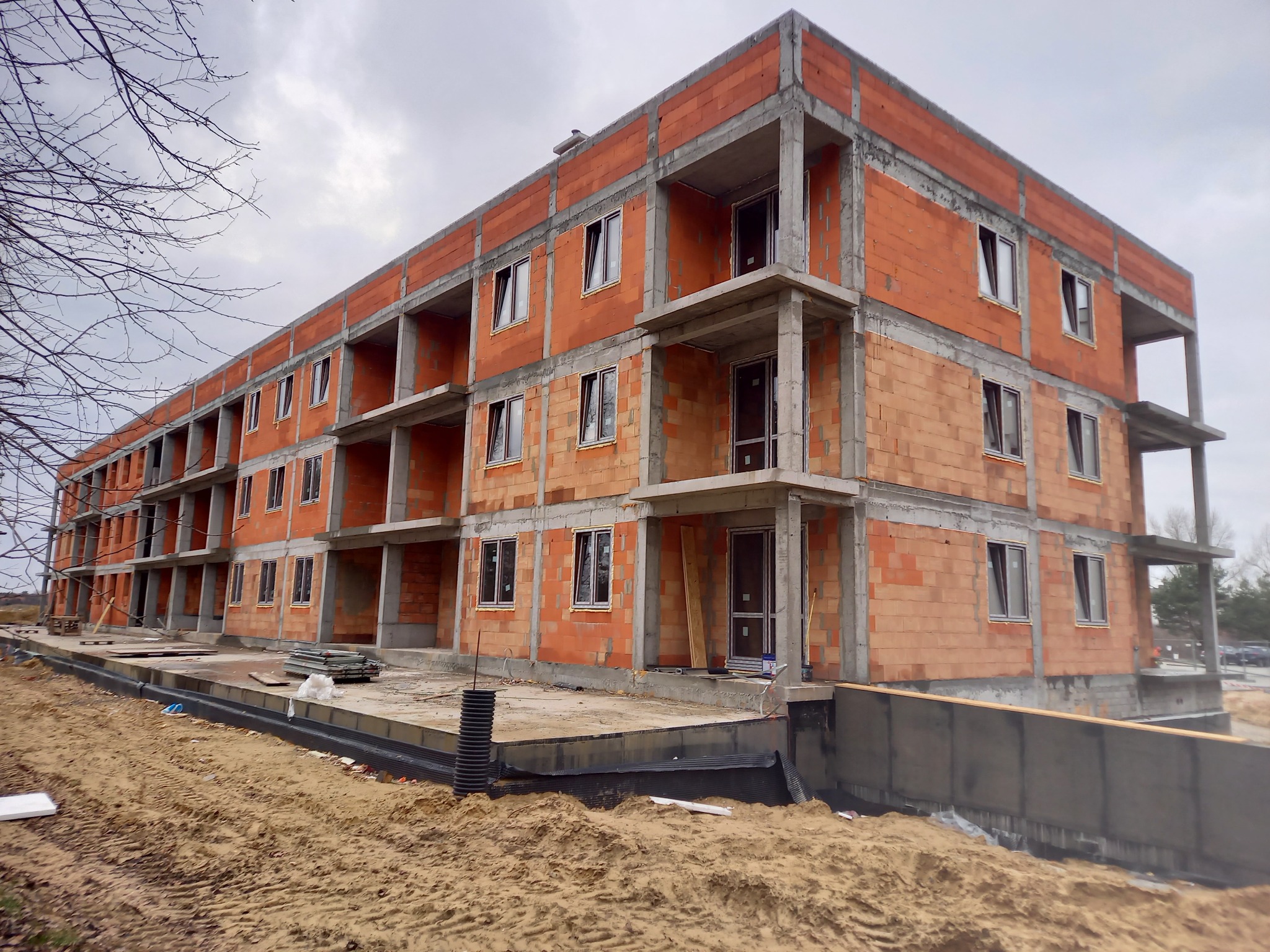 Trzykondygnacyjny budynek w stanie surowym otwartym z czerwonej cegły i betonu, widoczny z poziomu gruntu, z elementami izolacji fundamentów.