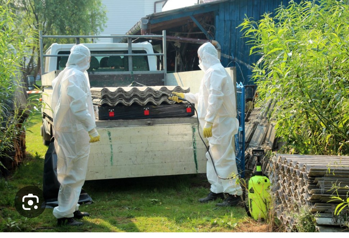 Pracownicy w kombinezonach zabezpieczających przygotowują do utylizacji płyty eternitowe na pace samochodu, na tle zieleni i zabudowań gospodarczych.