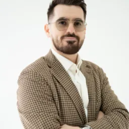 Portret mężczyzny w brązowej marynarce w pepitkę i okularach, z zegarkiem na ręce, na jasnym tle. Profesjonalny wizerunek.