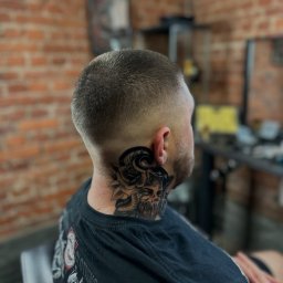 Under Zone Barber & Tattoo Studio Jelenia Góra - Krótko ostrzyżona męska głowa z tatuażem na szyi (róża i twarz). Widok z boku, na tle ceglanej ściany i wyposażenia salonu.