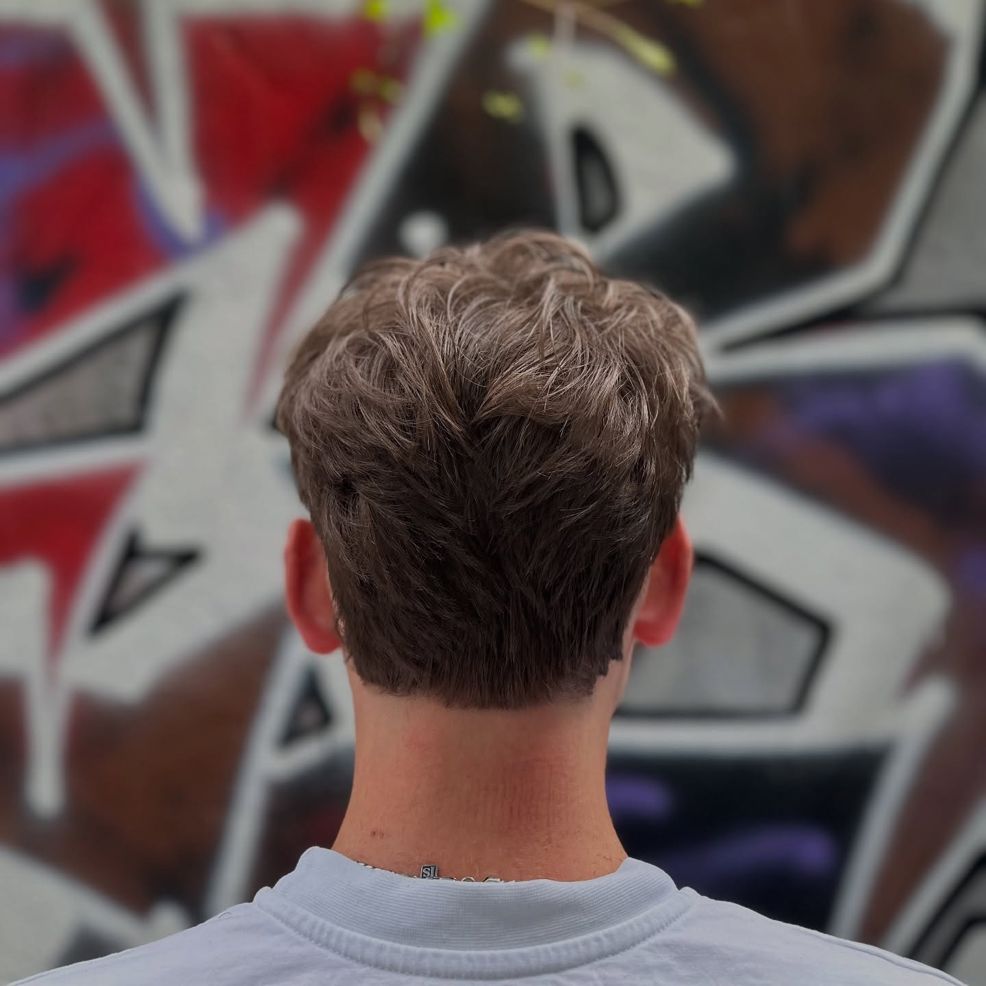 Tył głowy mężczyzny z modną, krótko ostrzyżoną fryzurą w odcieniach blondu i brązu, na tle ściany pokrytej kolorowym graffiti.