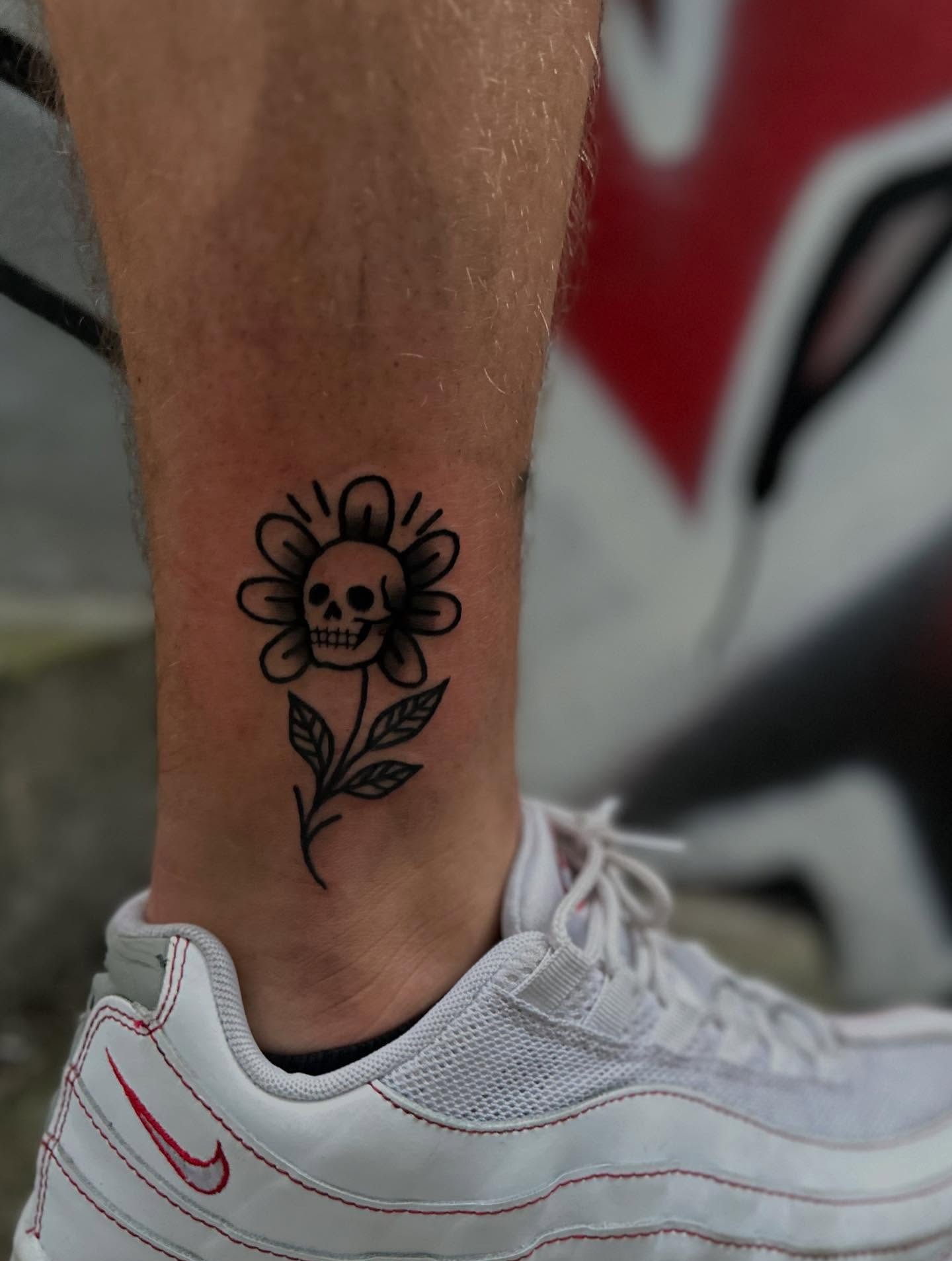 Czarno-biały tatuaż na kostce w Jeleniej Górze: kwiat z czaszką zamiast środka, widoczny fragment buta sportowego, tło w graffiti.