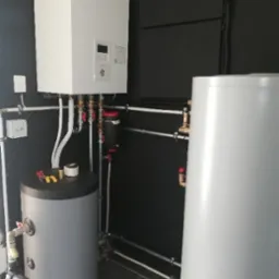 Biała pompa ciepła Panasonic z zbiornikiem na wodę, rury i armatura na tle ciemnej ściany. Widoczne narzędzia na zbiorniku, instalacja po montażu.