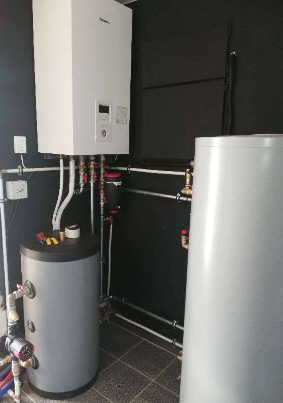 Biała pompa ciepła Panasonic z zbiornikiem na wodę, rury i armatura na tle ciemnej ściany. Widoczne narzędzia na zbiorniku, instalacja po montażu.